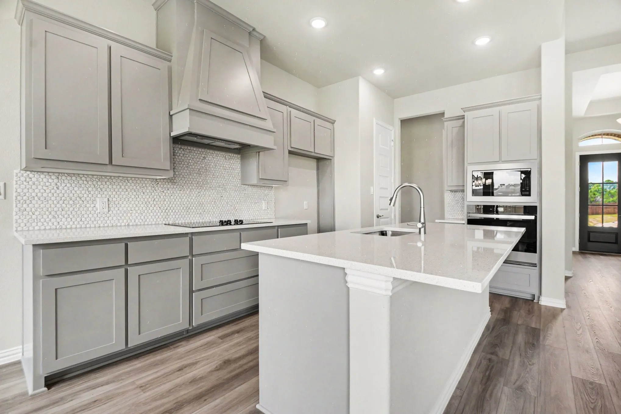 Kitchen featuring light gray cabinetry, white hexagonal tile backsplash, light-toned countertops, a central island with an undermount sink, and wood-finish flooring