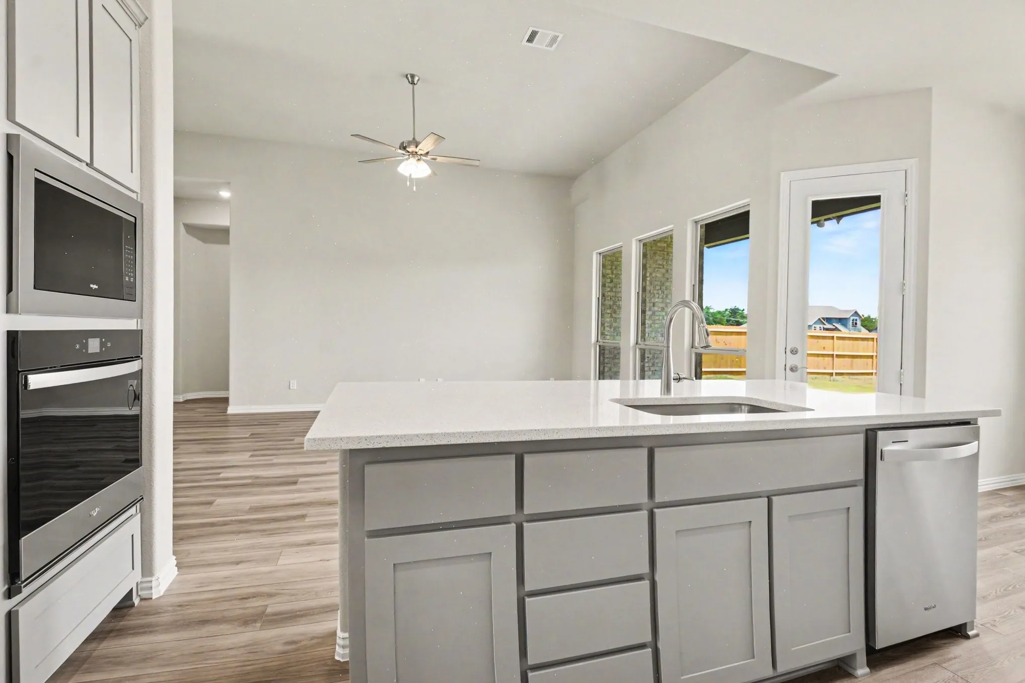 Open-concept kitchen featuring an island with a built-in sink and stainless steel dishwasher, light gray cabinetry, wood-finish flooring, and integrated stainless steel wall oven and microwave