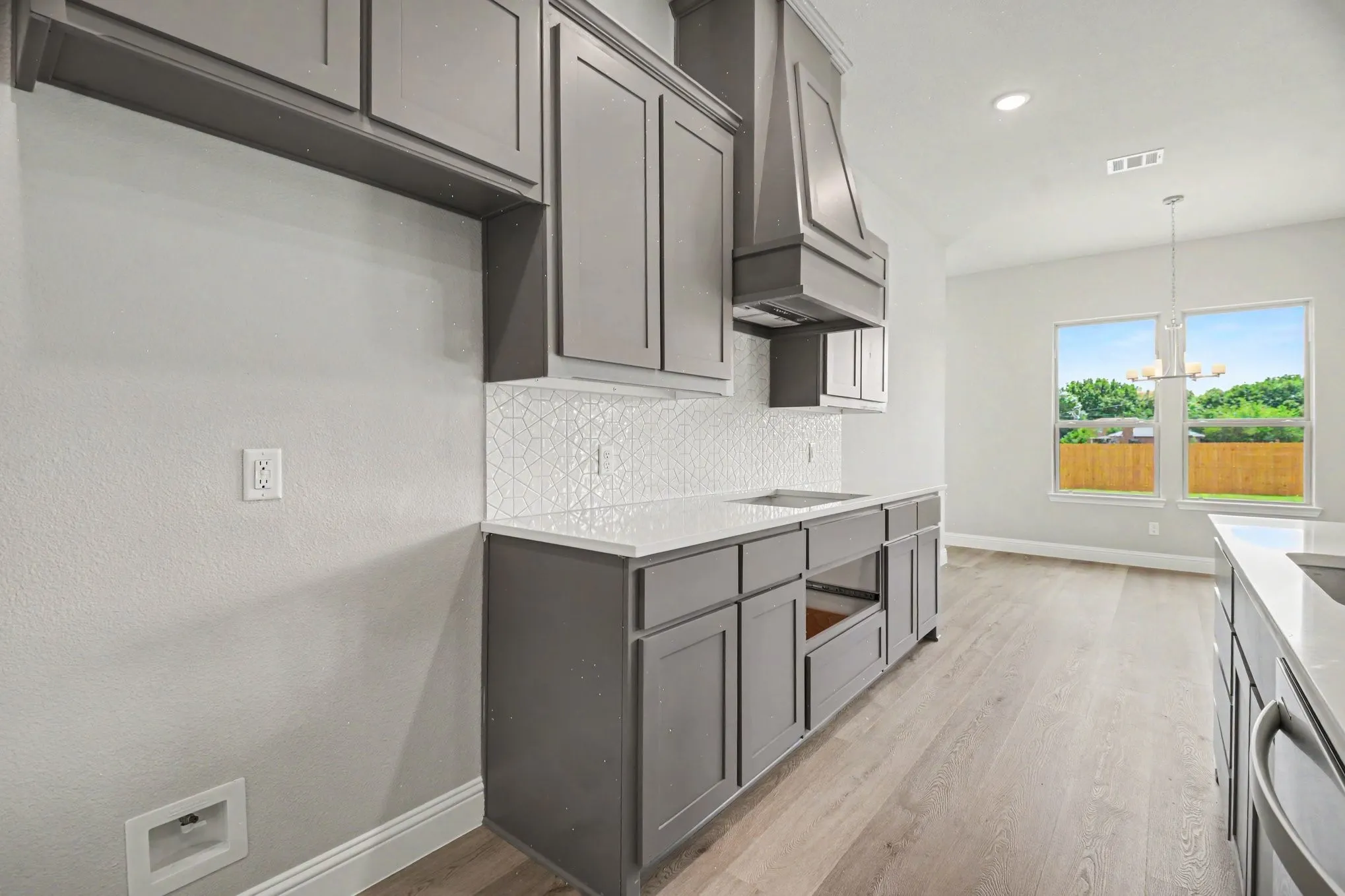 Contemporary kitchen featuring gray shaker cabinetry, white geometric tile backsplash, light-toned wood-finish flooring, and a stone countertop