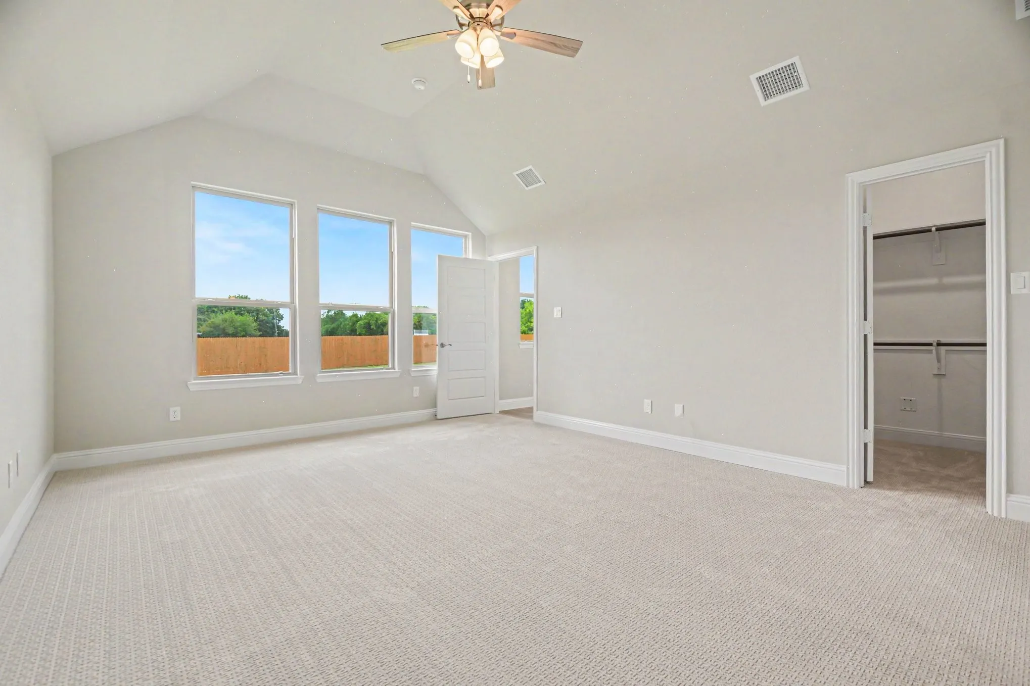 Spacious room featuring a vaulted ceiling with a ceiling fan, three large windows, textured carpet, and an open doorway revealing a closet with shelving and hanging rods