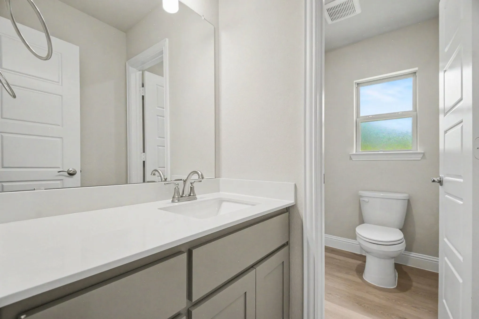 Bathroom featuring a light gray vanity with a white countertop and undermount sink, a large wall-mounted mirror, brushed nickel faucet, and wood-finish flooring