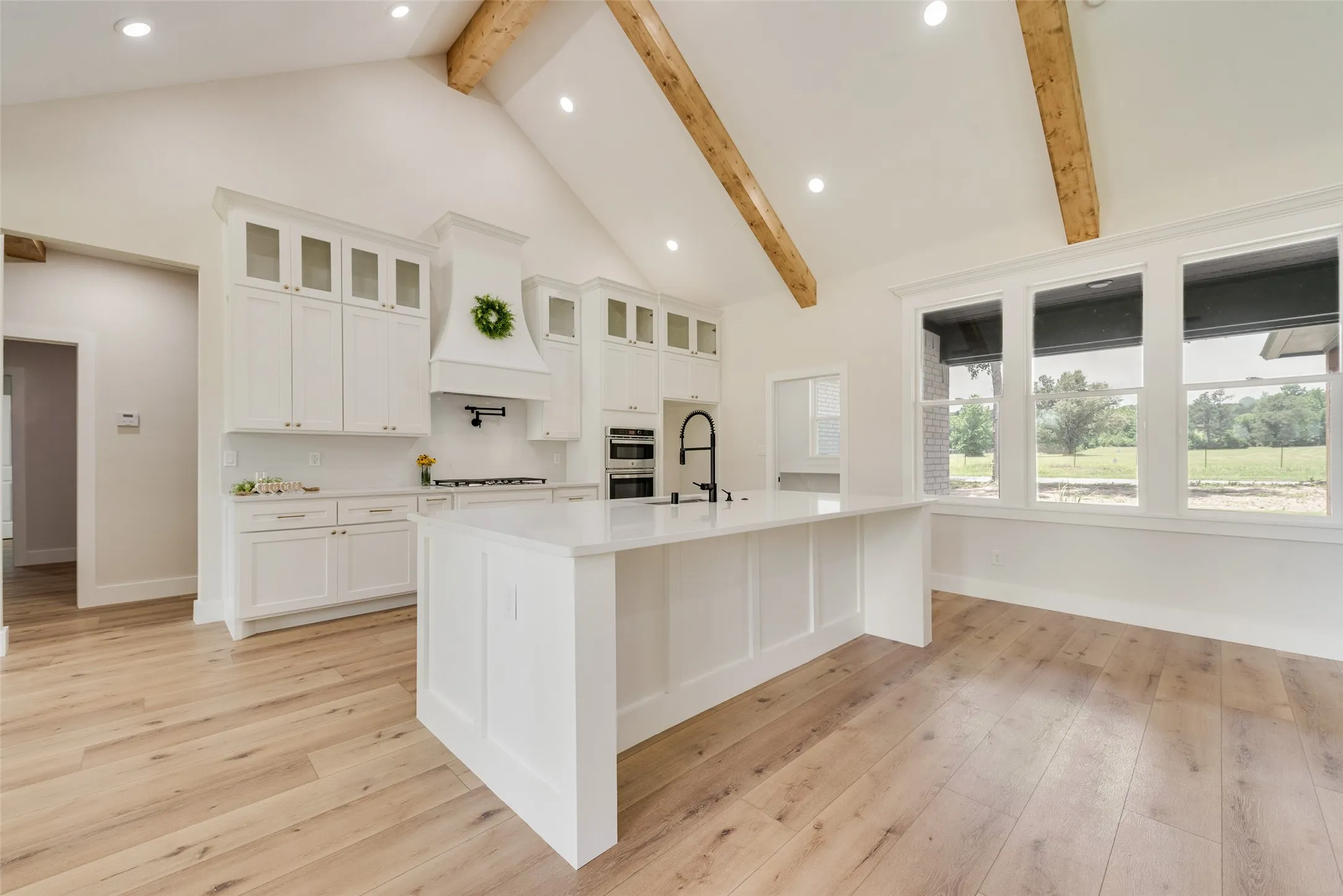 Bright kitchen featuring a large central island with a matte black faucet, white shaker cabinetry, wood-finish flooring, a vaulted ceiling with exposed wood beams, and a wall of windows