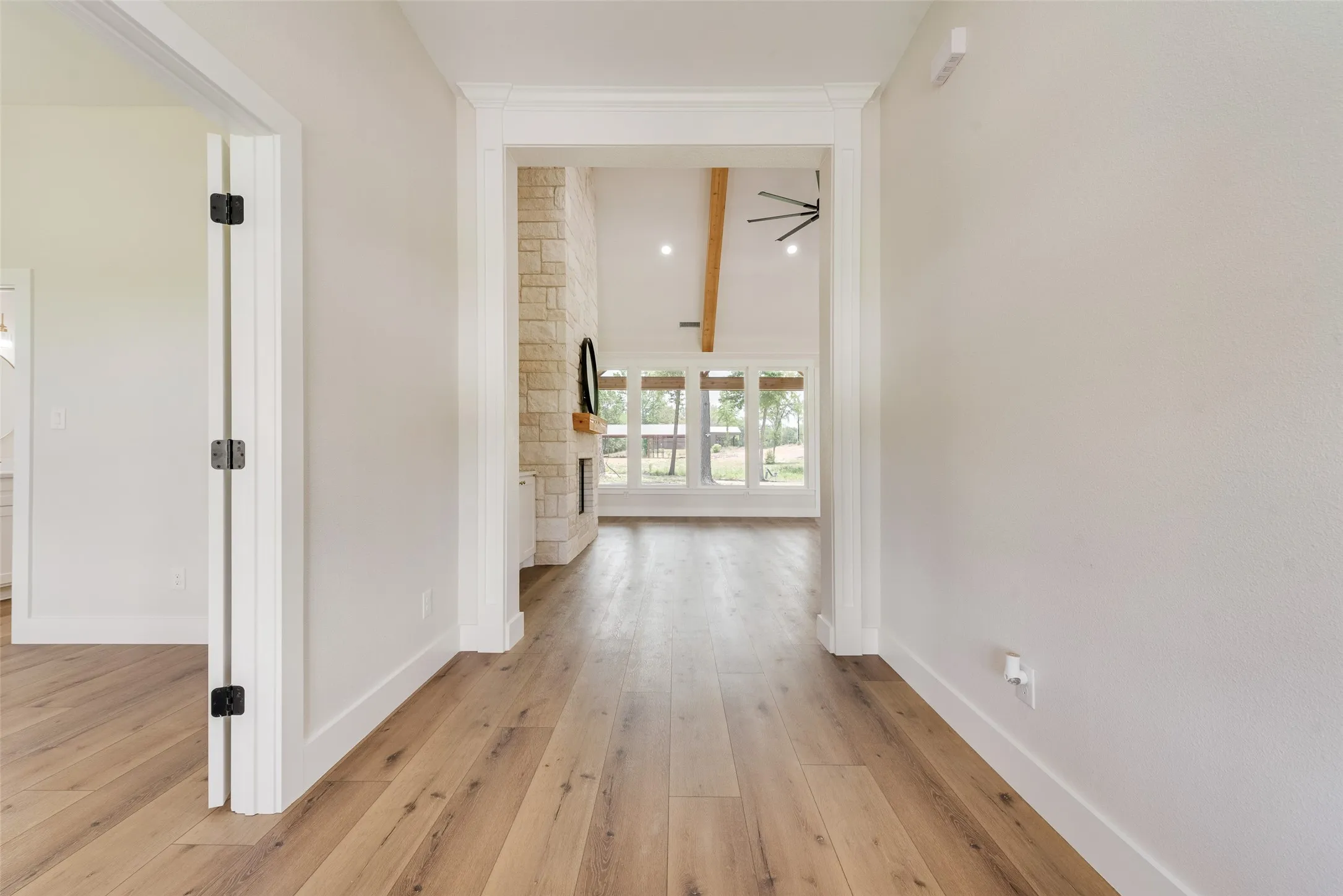 Hallway entry featuring wood-finish flooring, crisp white trim, and a clear sightline to a living space with a stone-clad fireplace, vaulted ceiling with exposed beams, and large picture windows