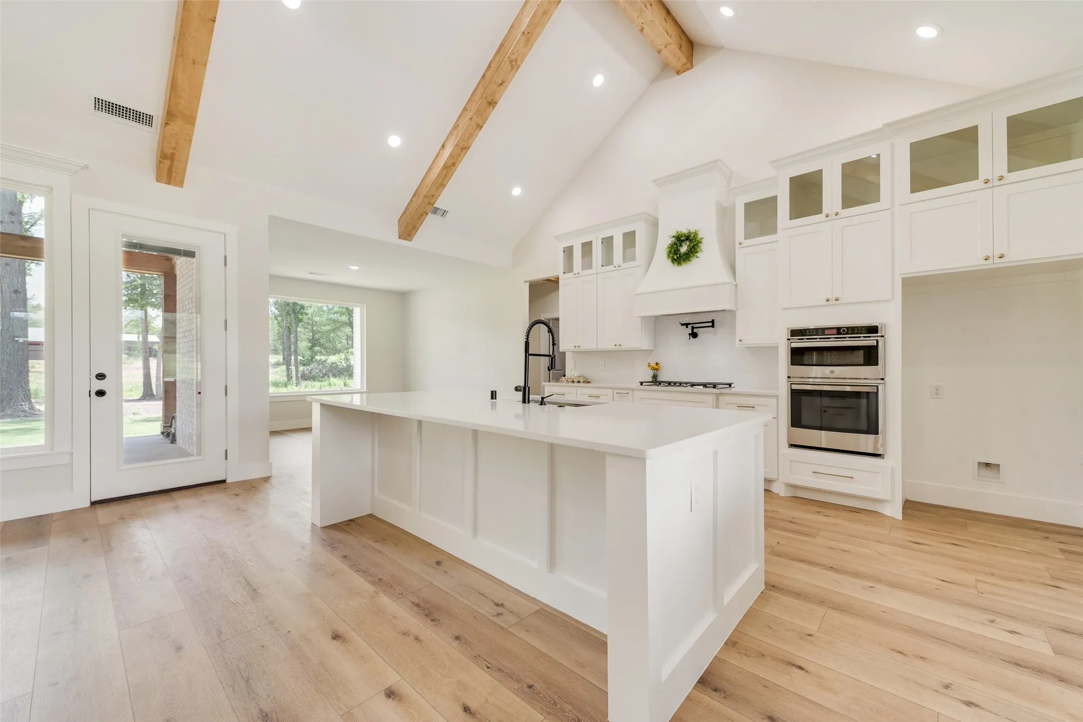 Spacious kitchen featuring a large central island with a solid surface countertop, white cabinetry with glass-front accents, and stainless steel built-in double ovens