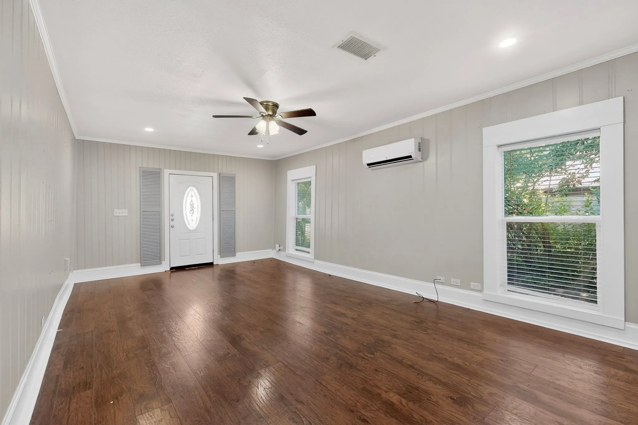 Spacious room featuring wood-finish flooring, light grey vertical paneling, and a white front entry door with an oval glass insert