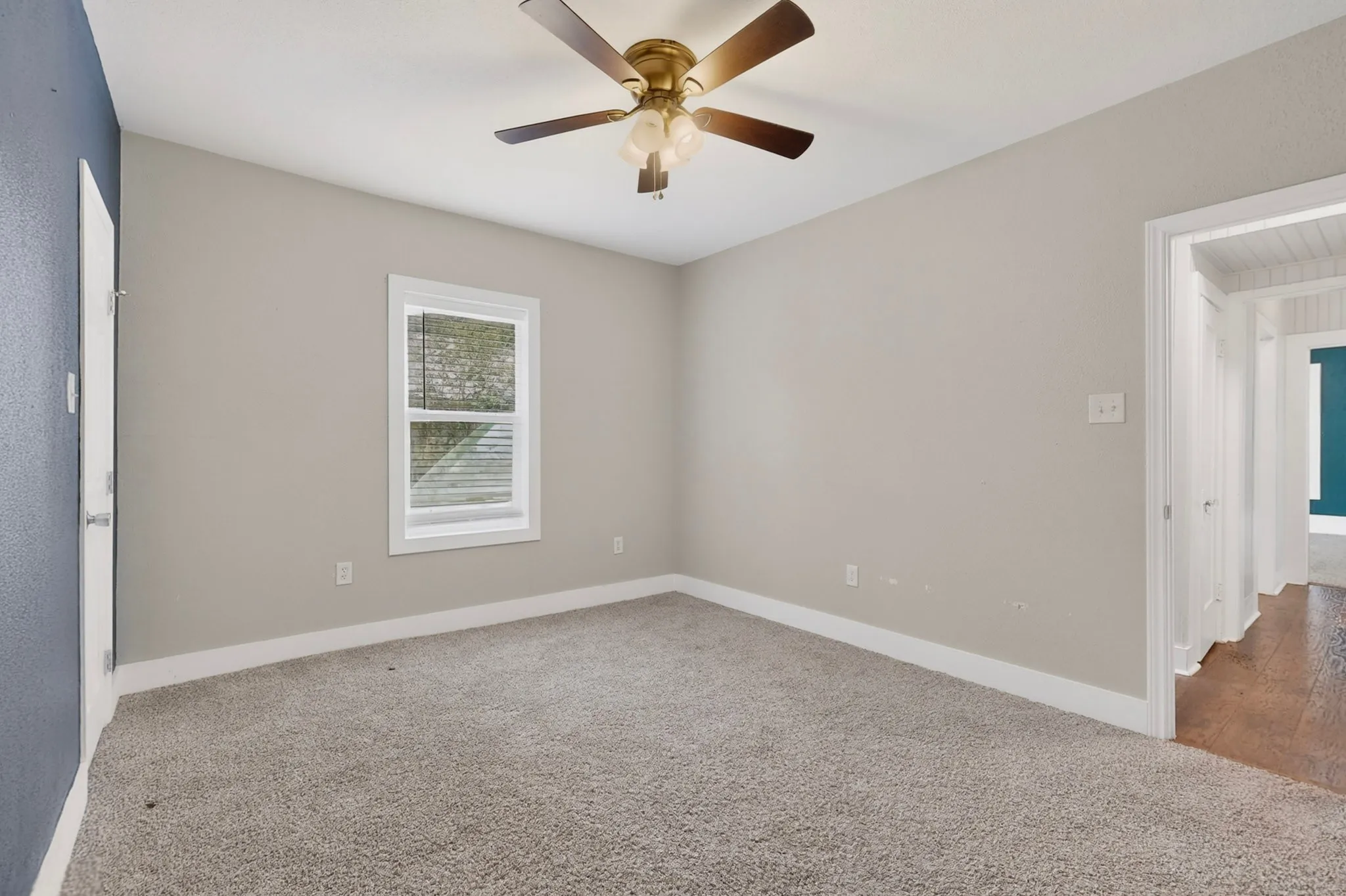 Carpeted room featuring a ceiling fan with light fixture, a window with white blinds, and a doorway opening to a wood-finish floor