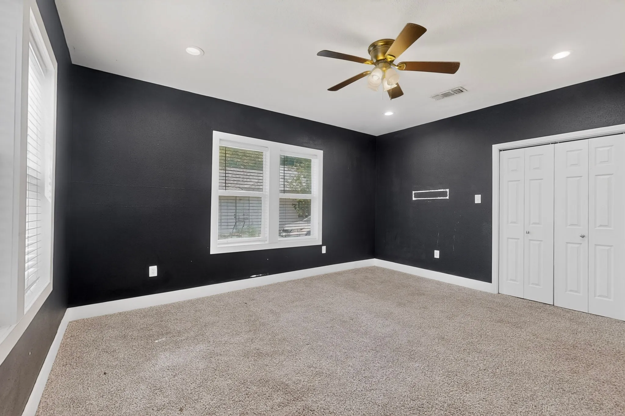 Spacious room featuring dark-painted walls, light-colored carpeting, two white-framed windows with blinds, a ceiling fan with integrated lighting, and white bi-fold closet doors