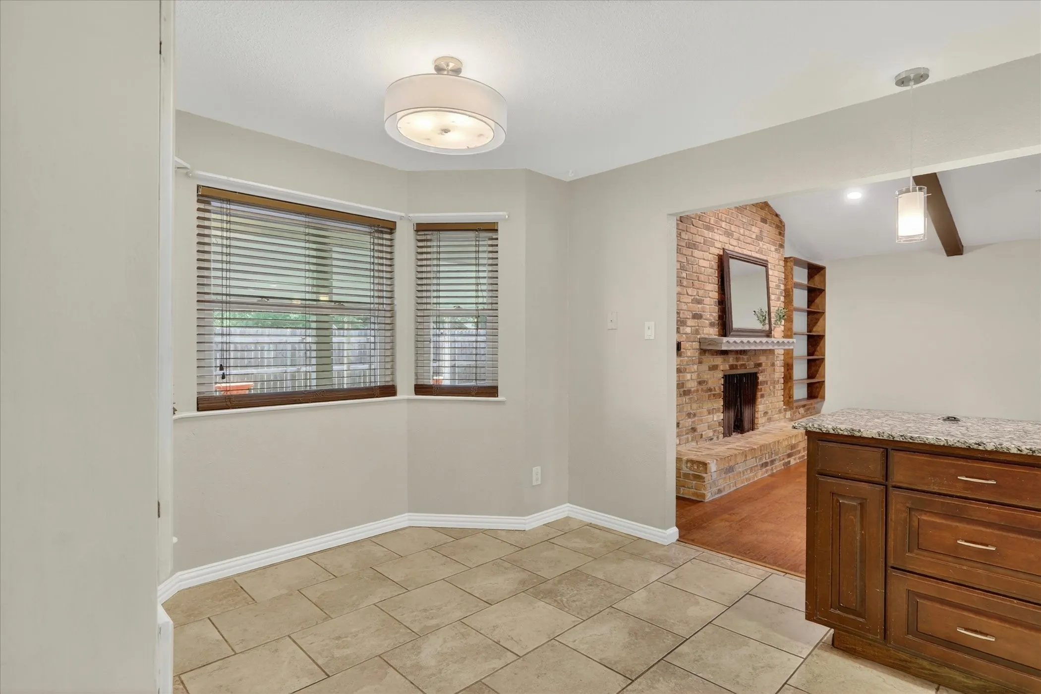 Light-colored tile flooring, breakfast nook with bay windows, wood-finish blinds, built-in cabinetry with stone countertop, exposed brick fireplace with wood mantel