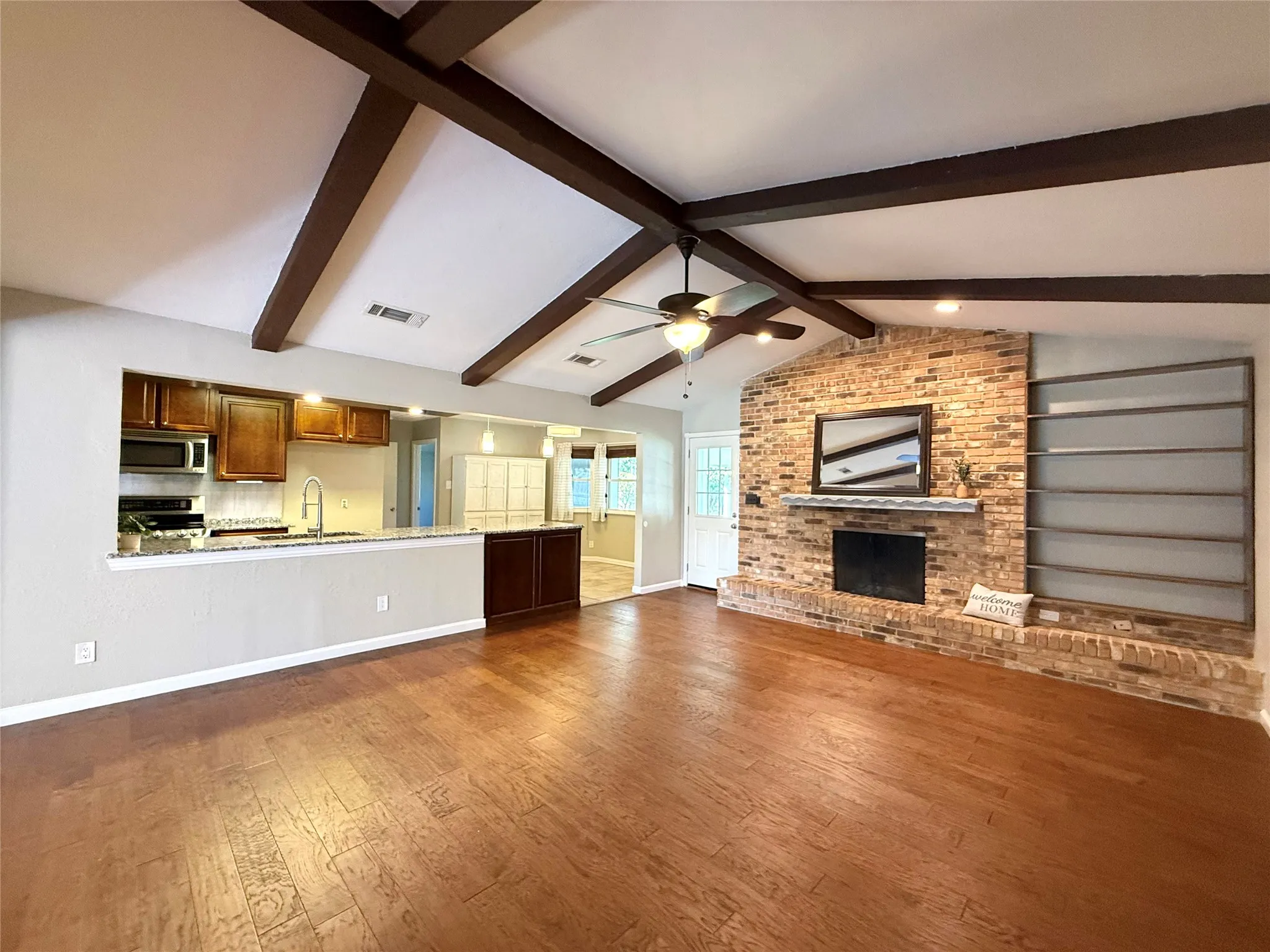 Expansive room featuring wood-finish flooring, exposed ceiling beams, a brick fireplace, and built-in shelving