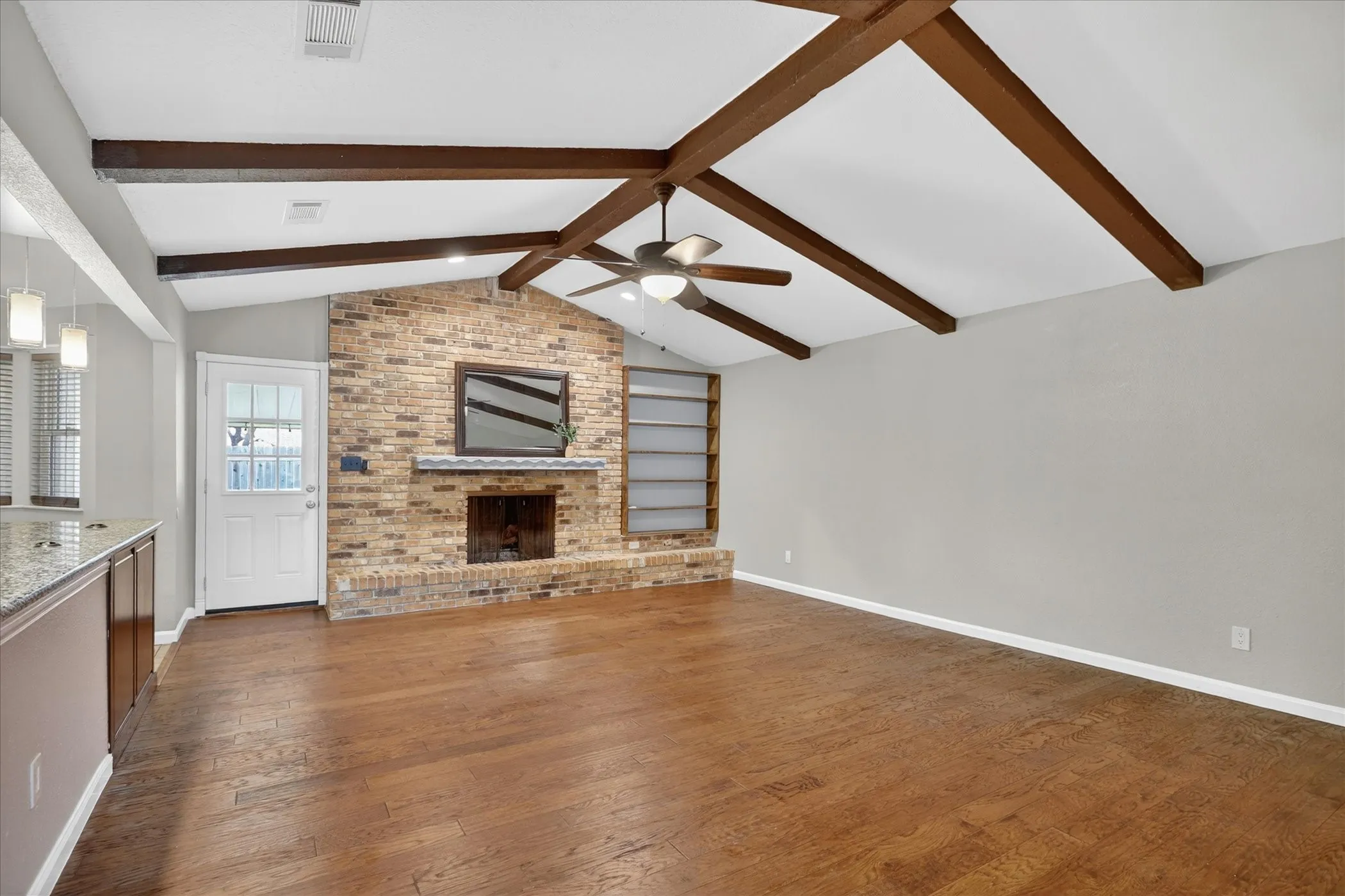 Expansive room featuring wood-finish flooring, exposed ceiling beams, a brick fireplace, and built-in shelving