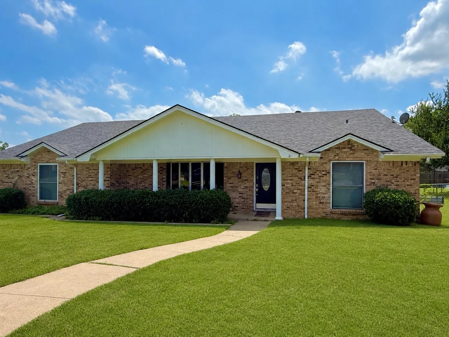 Traditional brick facade featuring a covered front porch with white columns, a dark entry door with oval glass insert, and a gabled roofline. Grass enhanced with AI.