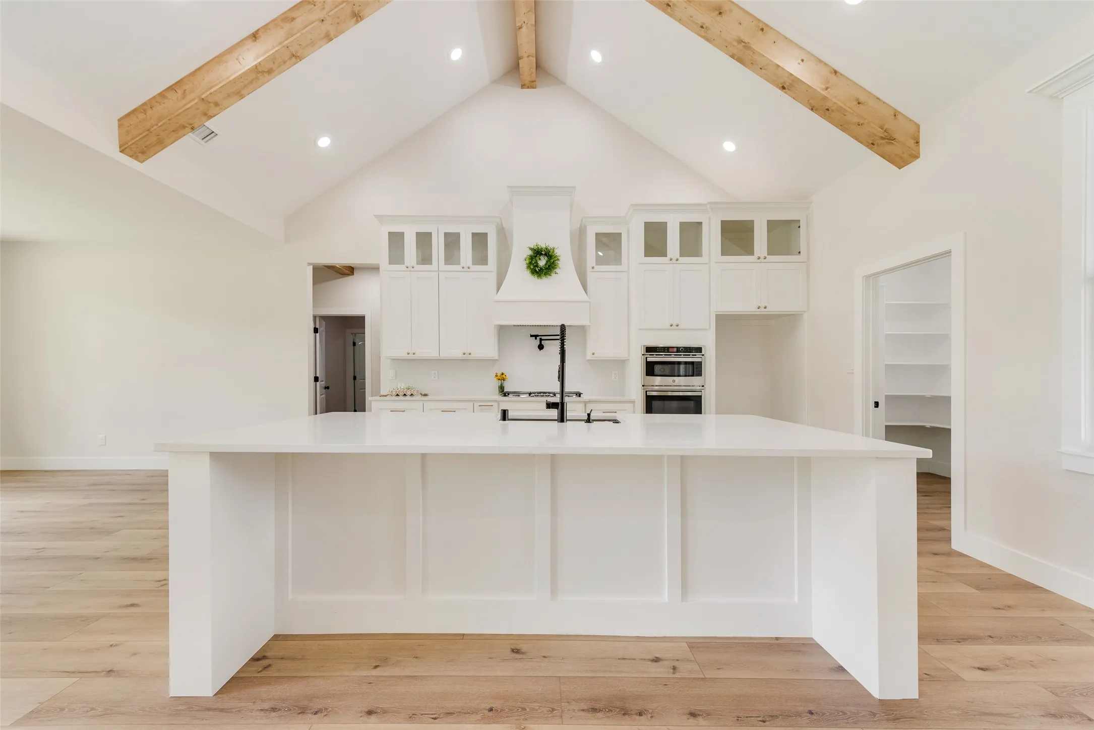 Gourmet kitchen featuring a large central island with white paneling, vaulted ceiling with exposed wood beams, white shaker cabinetry, integrated double ovens, and wood-finish flooring