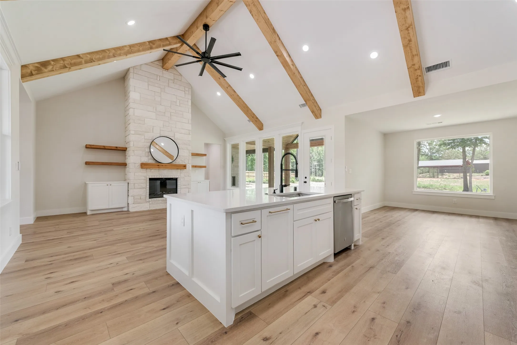 Open-concept living area featuring a vaulted ceiling with exposed wood beams, a stone-clad fireplace, and wood-finish flooring