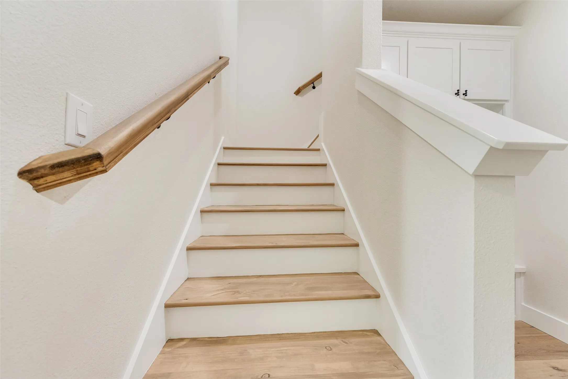 Staircase featuring wood-finish treads, white risers, a natural wood handrail, and a white newel post with cap