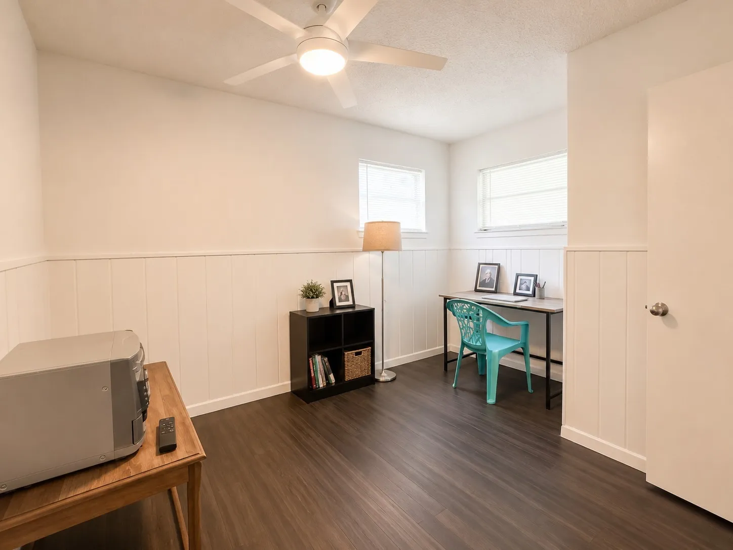 Room featuring dark wood-finish flooring, white wainscoting, two windows, and an overhead ceiling fan with lighting