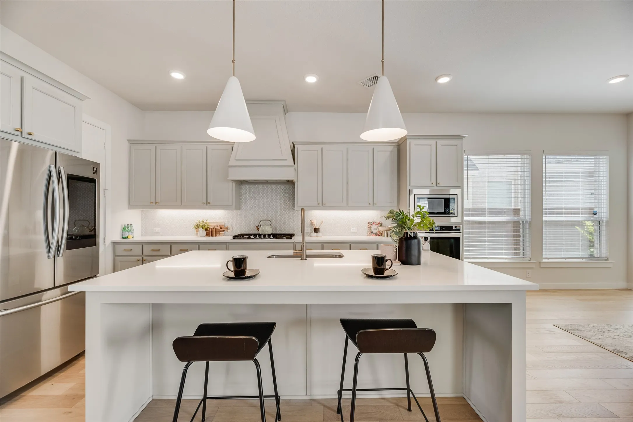 Modern kitchen featuring an expansive island with a waterfall edge, stainless steel appliances, light-toned cabinetry, two pendant lights, and recessed ceiling lights