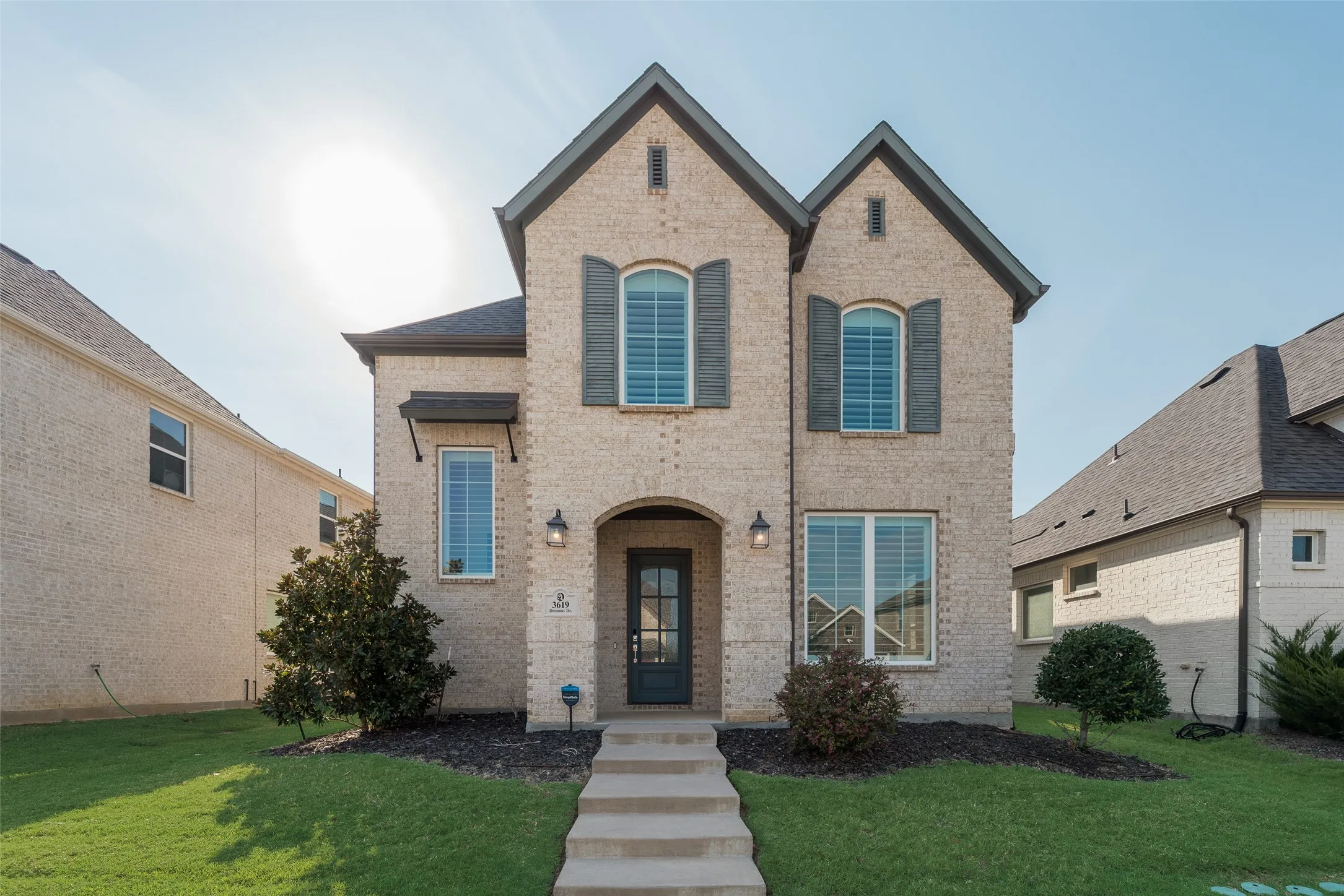 Two-story brick facade residence featuring dark shutters, arched entryway, a neatly maintained front lawn, and a paved walkway