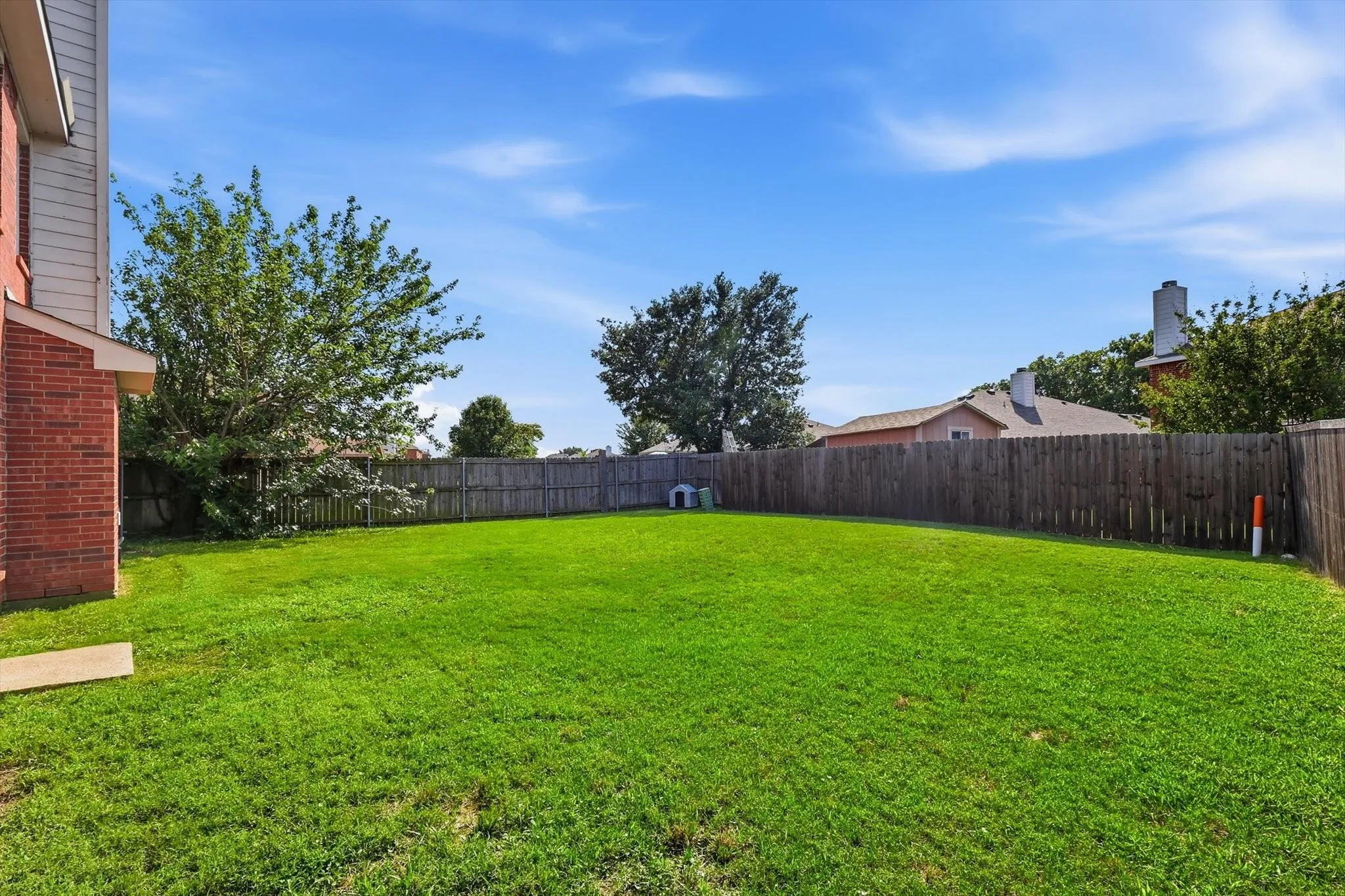 Expansive grassy backyard featuring a full privacy fence, mature trees, and a brick exterior wall
