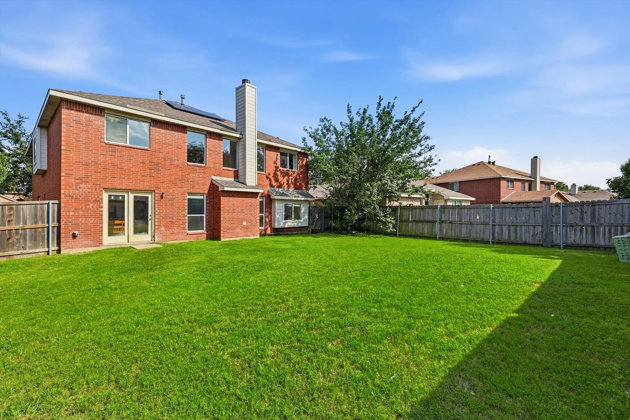Expansive fenced backyard featuring a manicured lawn, red brick exterior, dual-pane windows, double glass doors, and a prominent chimney structure