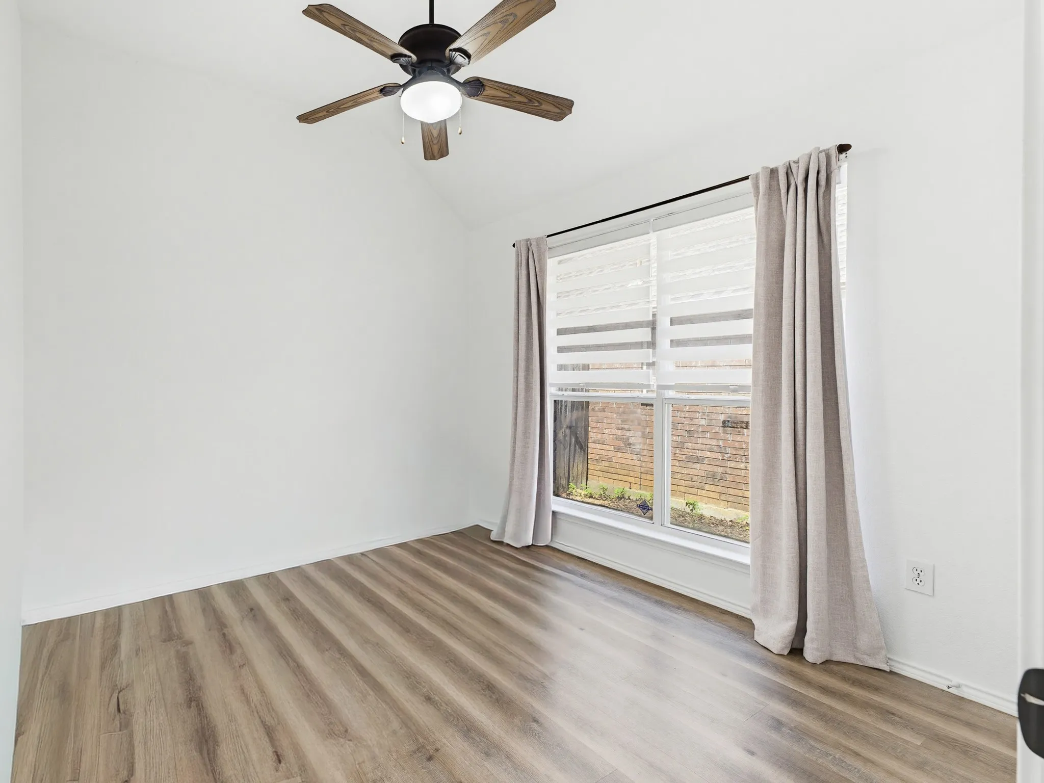 Room featuring wood-finish flooring, a vaulted ceiling, a ceiling fan with integrated lighting, and a large window with blinds and drapes