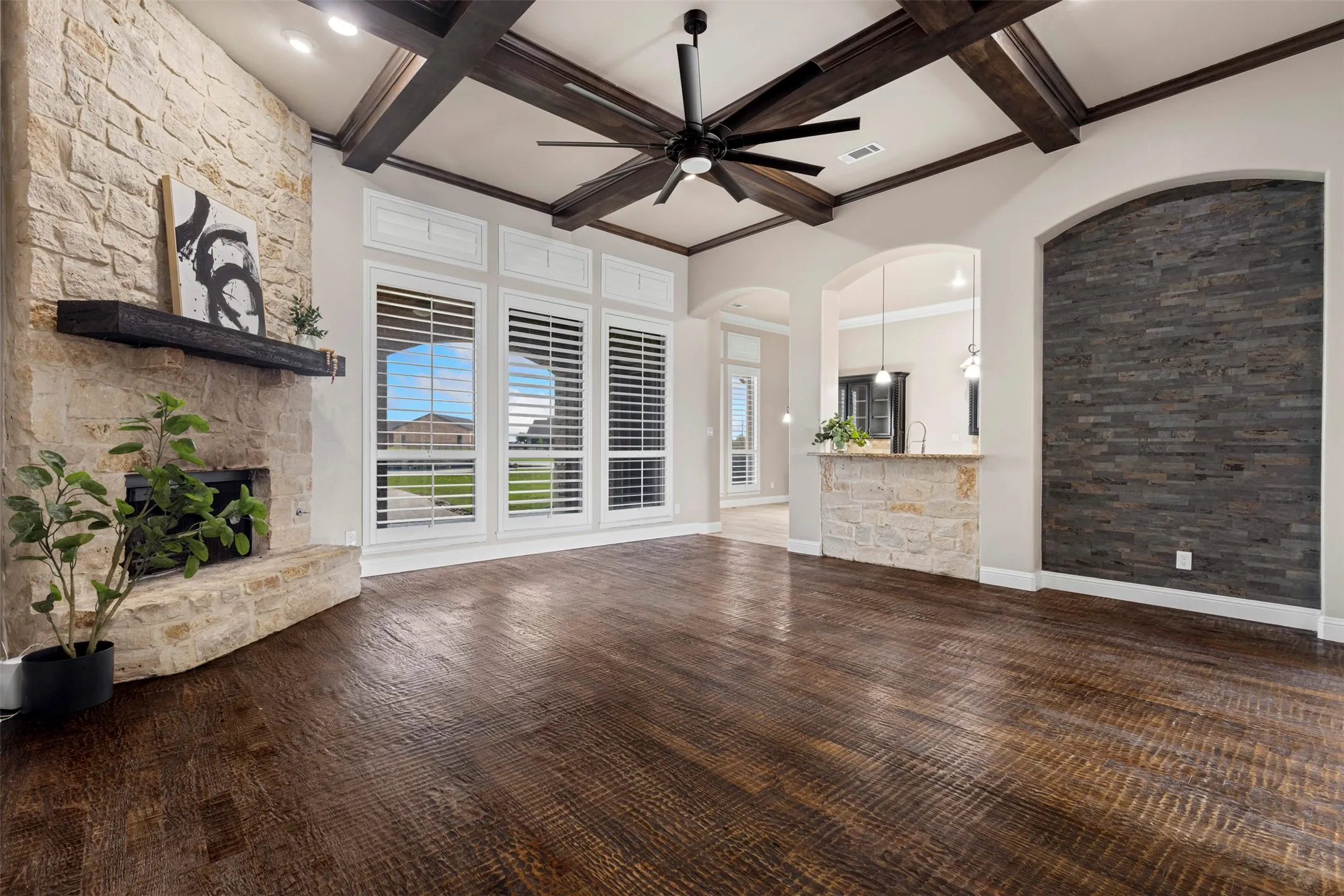Spacious living area featuring dark wood-finish flooring, a stone-clad fireplace with a dark wood mantel, and large windows with plantation shutters