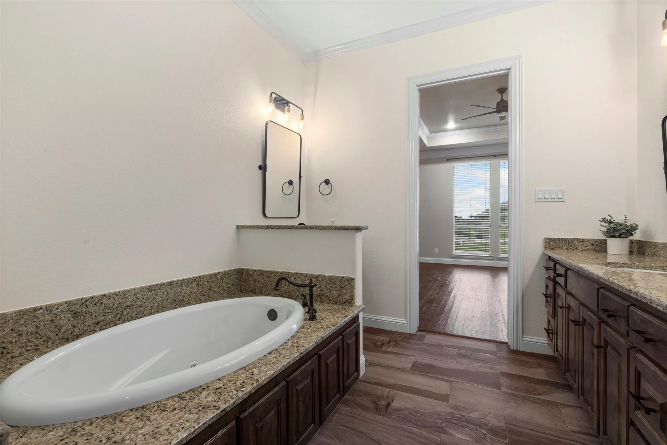 Built-in soaking tub with granite surround, dark wood cabinetry, dual vanity with granite countertop, and wood-finish flooring
