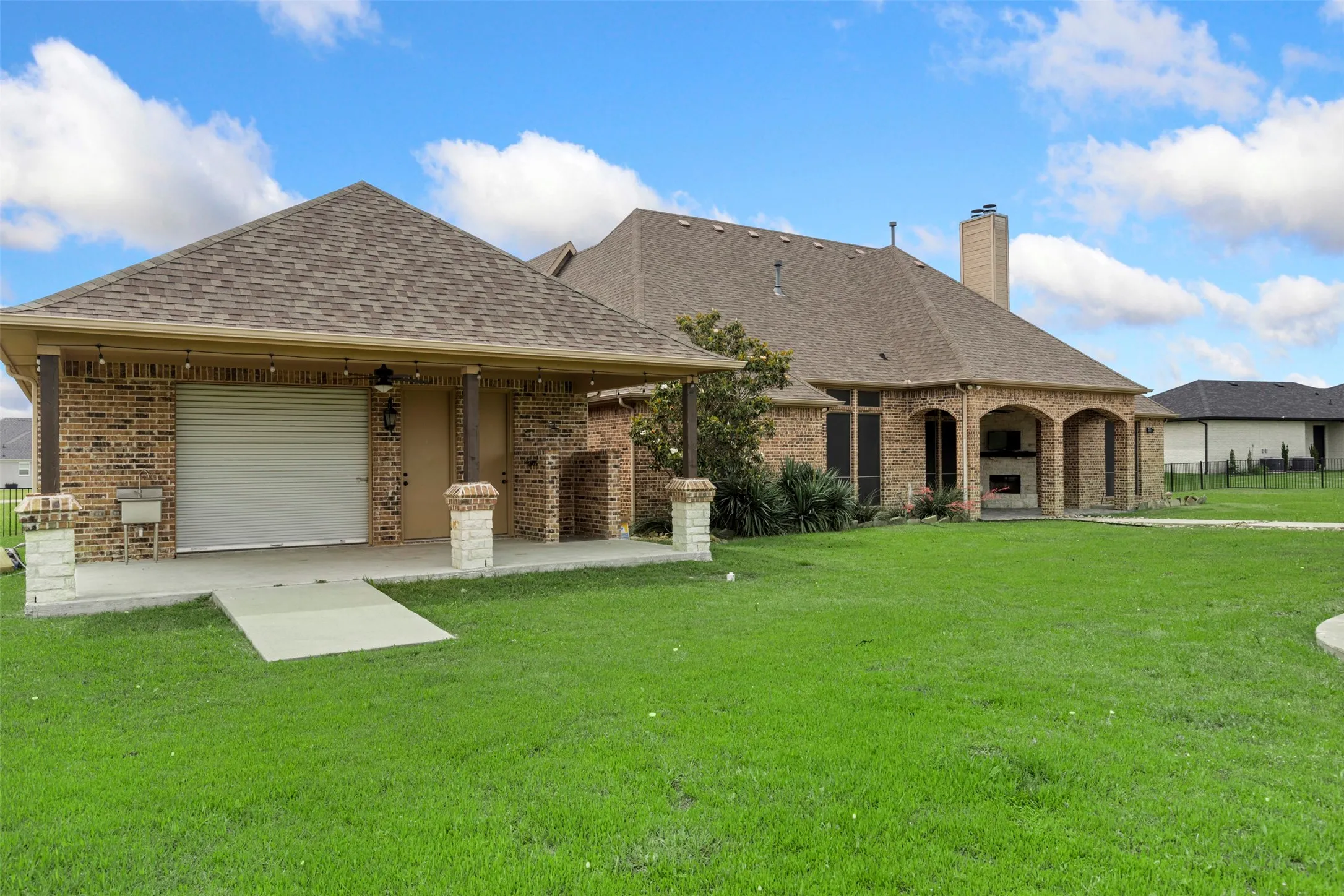 Expansive green lawn with a detached brick outbuilding featuring a roll-up door, outdoor sink, and covered patio with string lighting