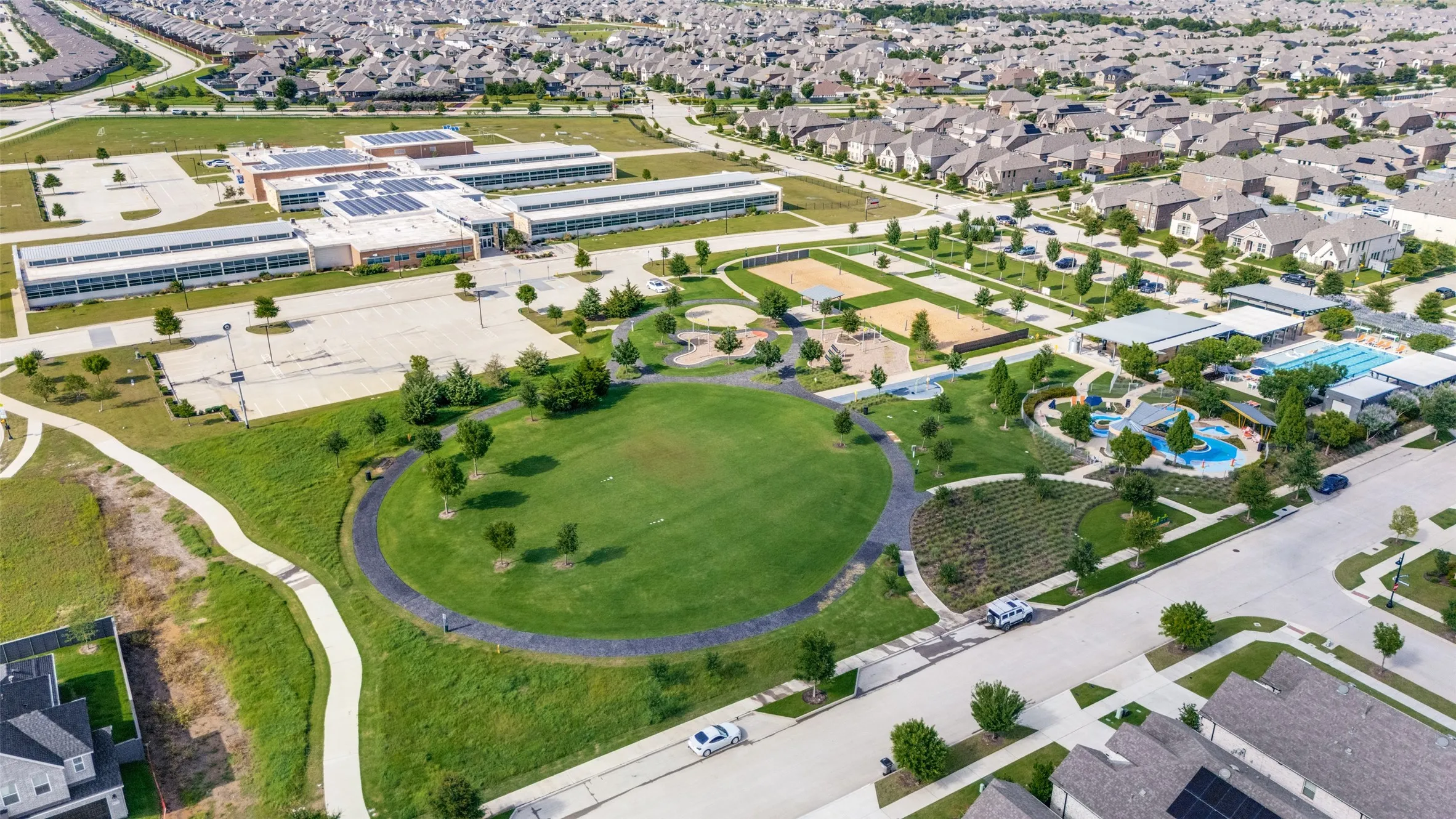 Expansive aerial view showcasing a neighborhood with a large community building featuring solar panels, multiple recreational swimming pools, a central green space, and extensive paved pathways