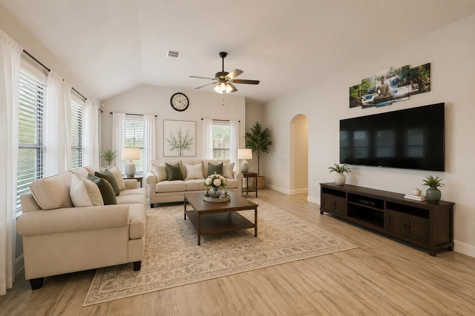 Spacious living area featuring vaulted ceilings, wood-finish flooring, multiple windows with blinds and sheer curtains, a ceiling fan with light fixture, and a built-in media console