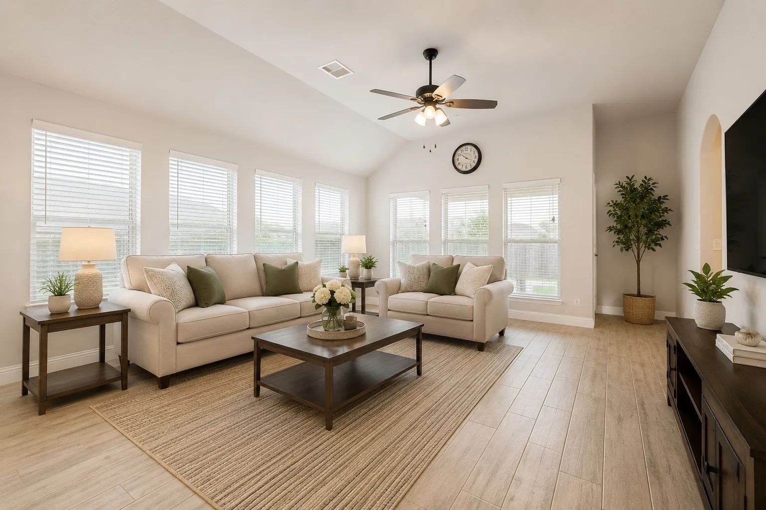 Spacious living area featuring light wood-finish flooring, vaulted ceilings with a ceiling fan, and multiple windows providing natural illumination