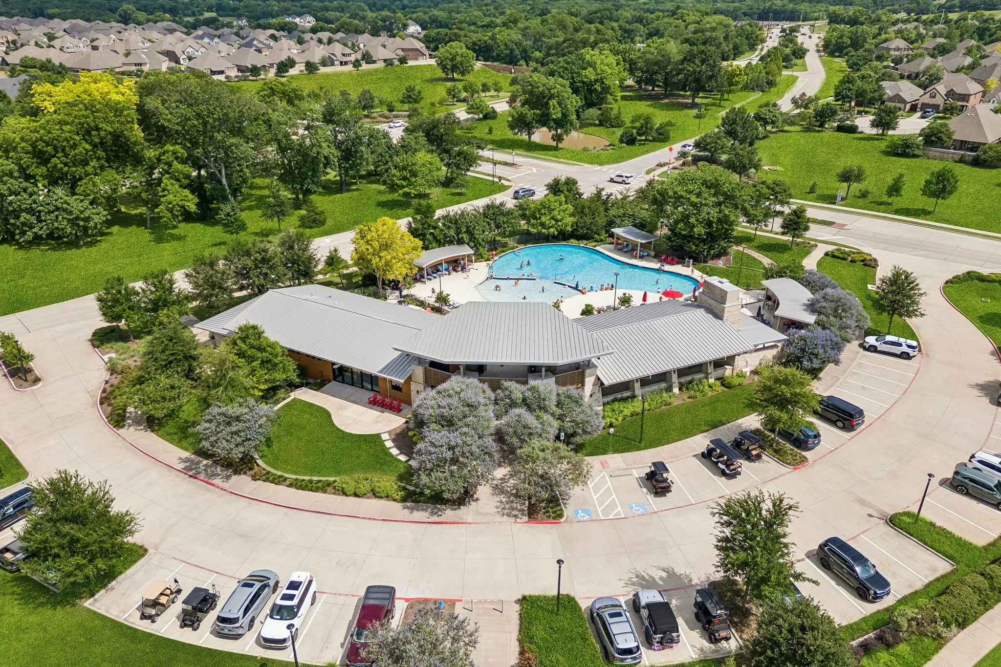 Community amenity center featuring a large swimming pool, expansive parking areas, and surrounding green spaces
