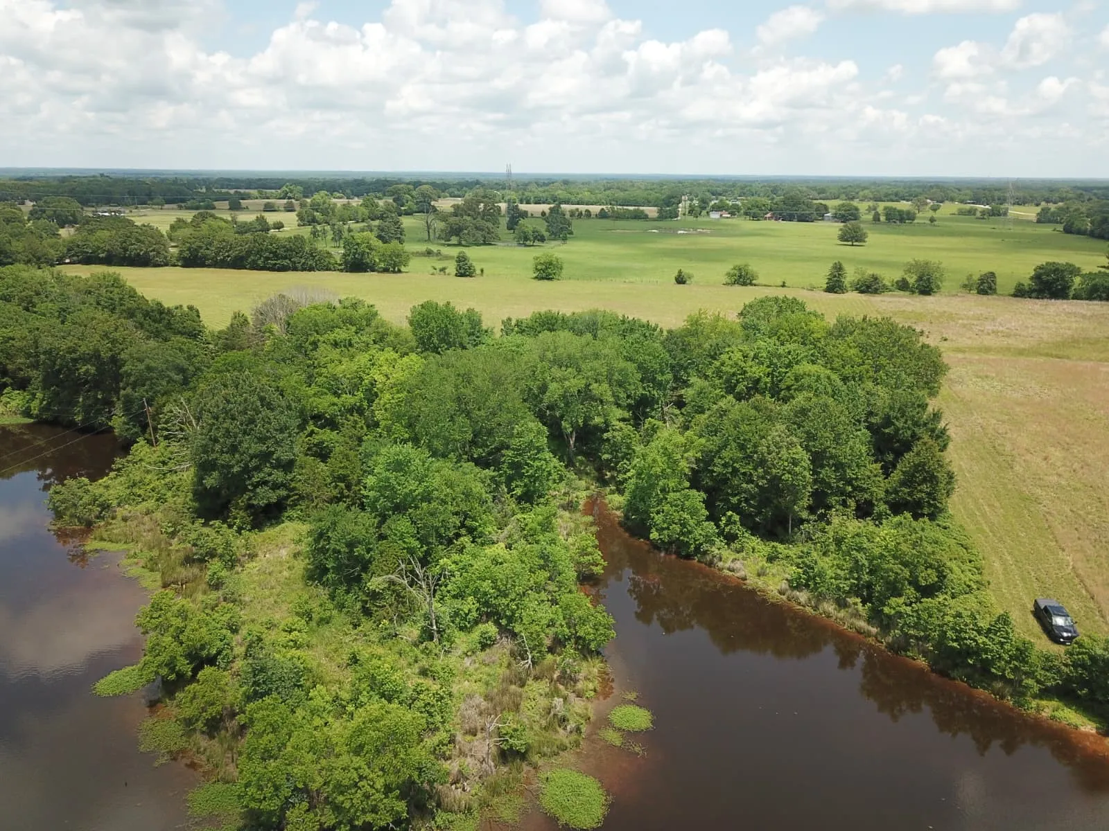 Expansive natural landscape featuring a substantial body of water, mature tree lines, and open fields under a spacious sky