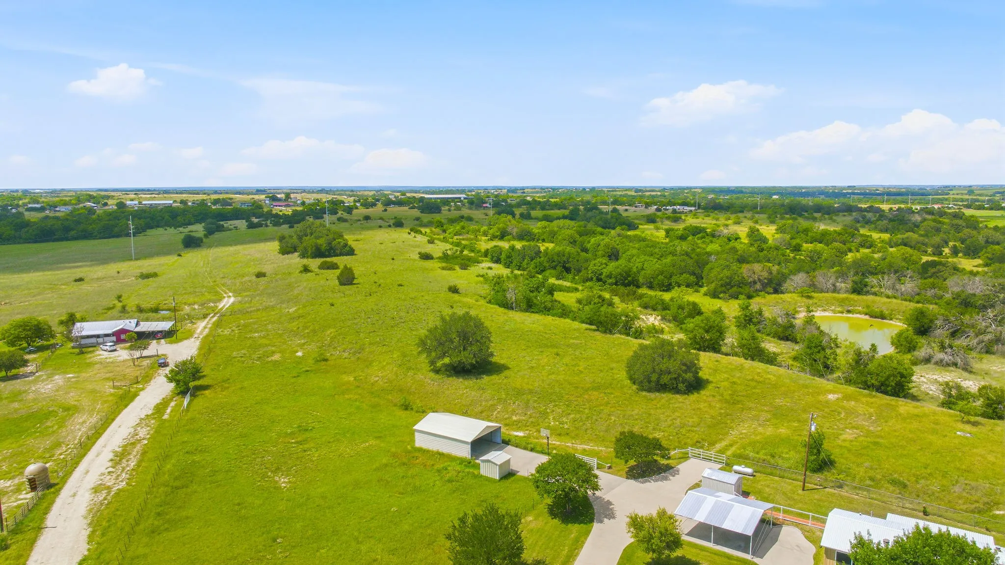Expansive green acreage featuring a detached red-roofed structure, several outbuildings, paved driveways, and a natural pond