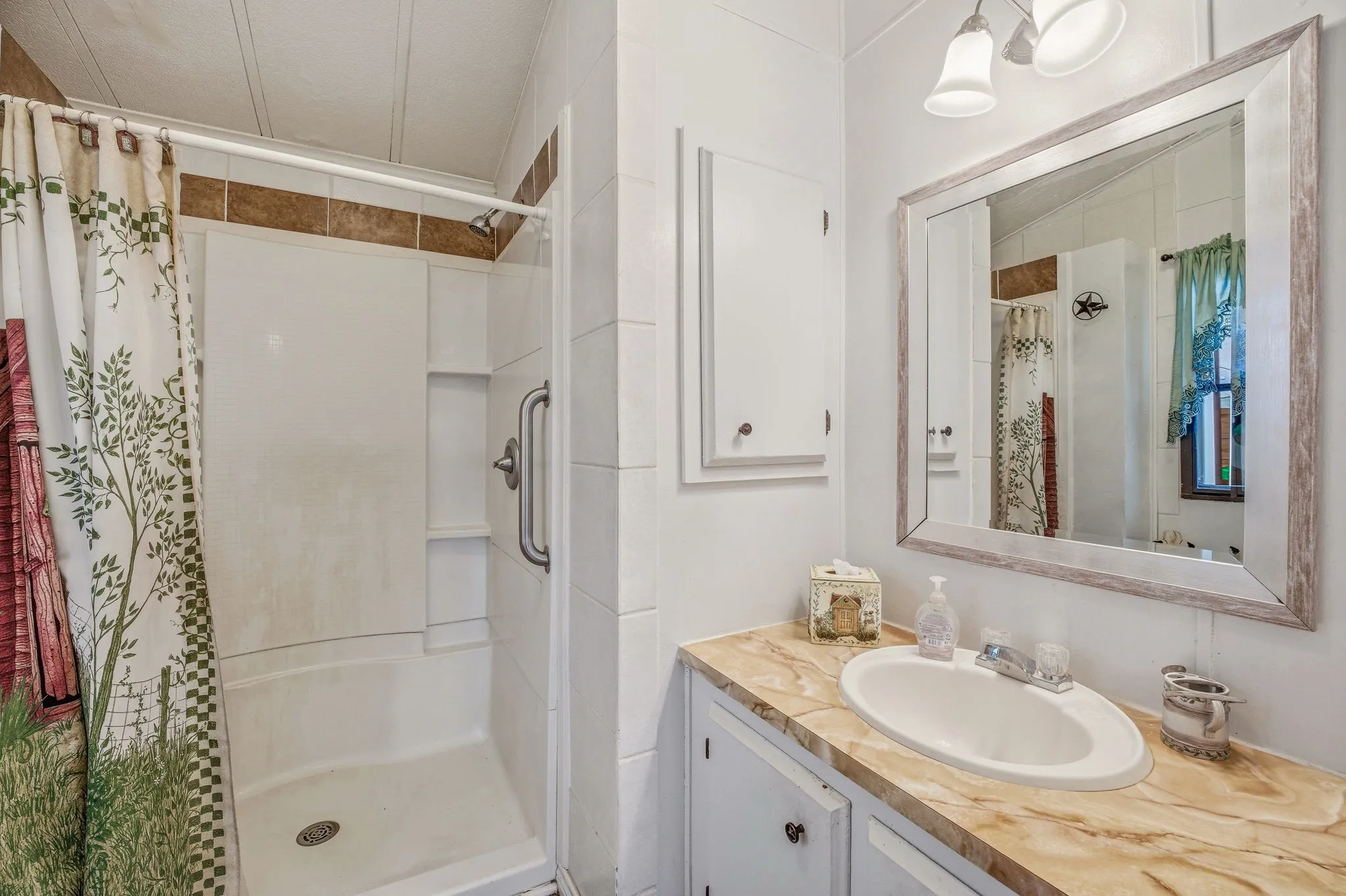 Bathroom featuring a white shower stall with a grab bar, a single vanity with an oval sink, a light-colored patterned countertop, and a framed mirror with an overhead light fixture