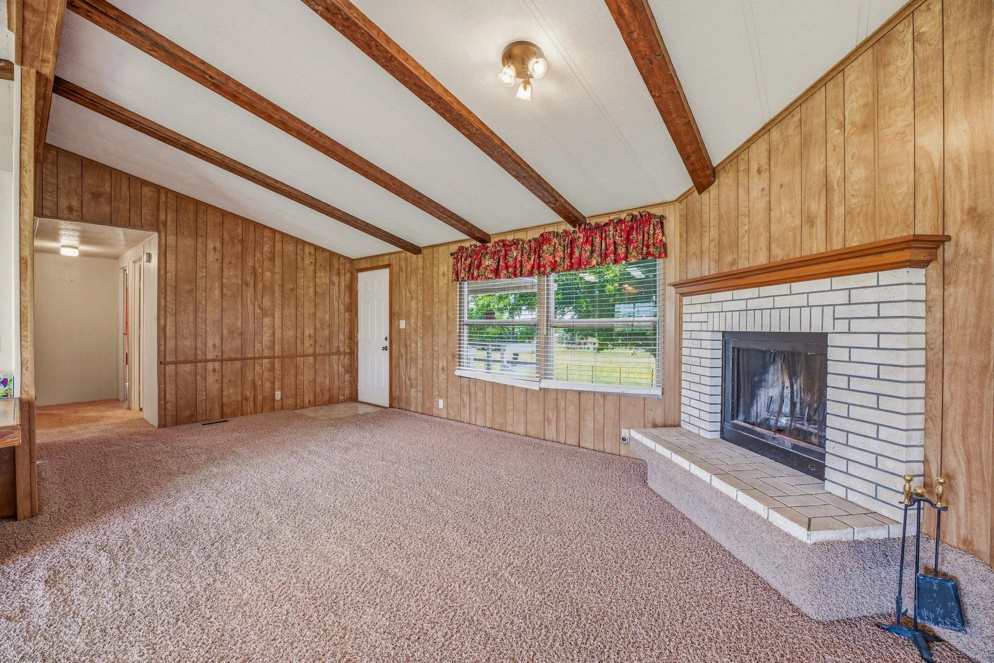 Spacious room featuring a white brick fireplace with a wood mantle, wood-paneled walls, exposed wood ceiling beams, and light brown carpeting