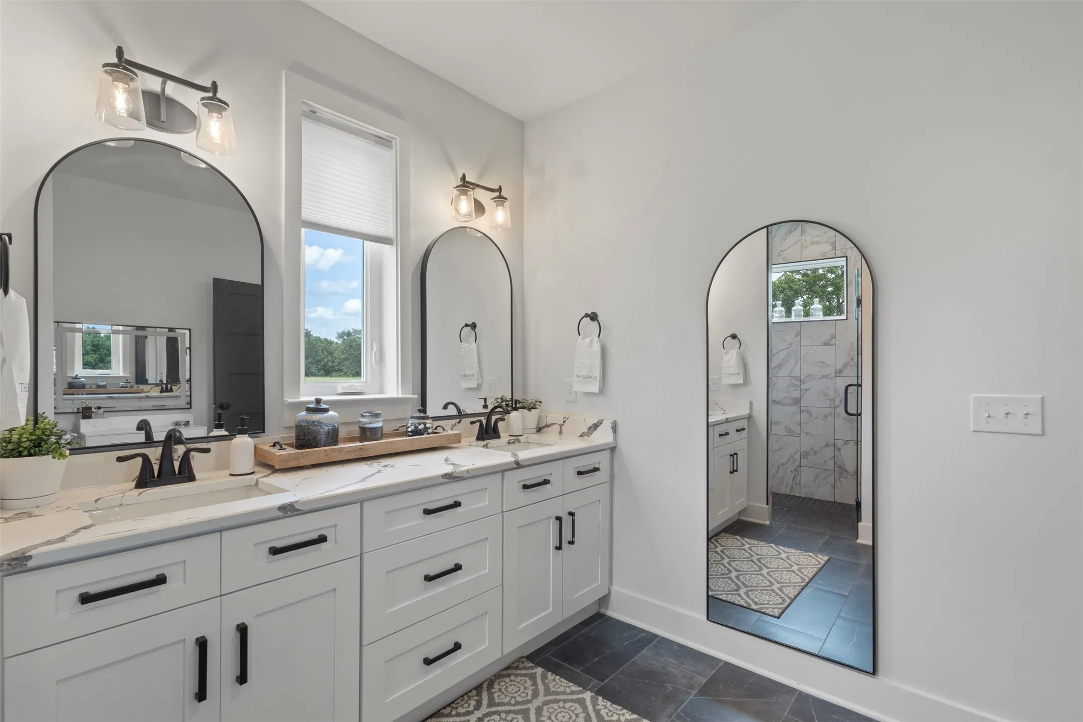 Spa-inspired primary bath featuring dual vanities, designer lighting, sleek black hardware, and a spacious walk-in shower with refined tilework—creating a polished, luxury retreat with modern country elegance.