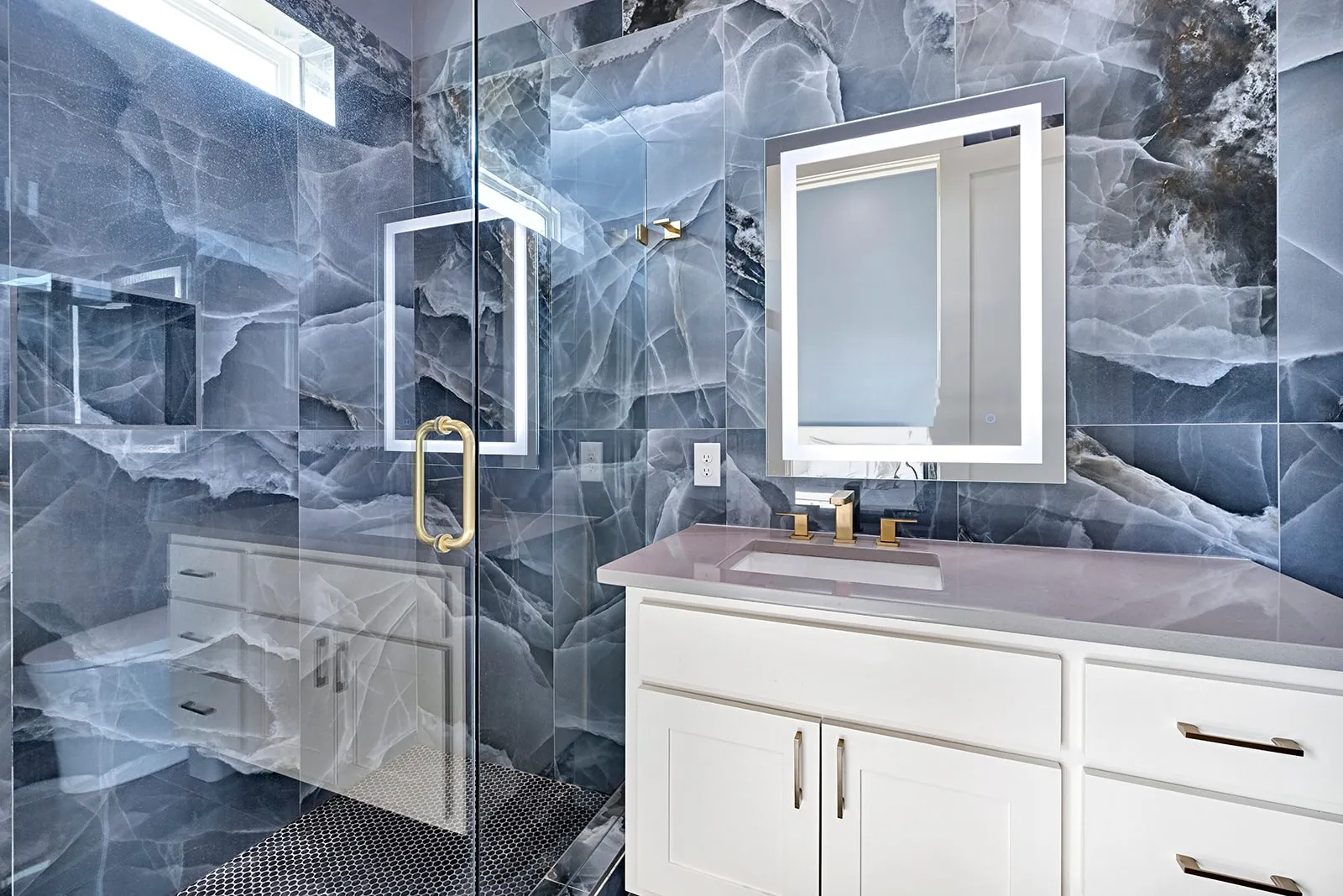 Spacious bathroom featuring full-height stone-look tile walls, a frameless glass shower enclosure with a gold-tone handle, and a white vanity with a light countertop