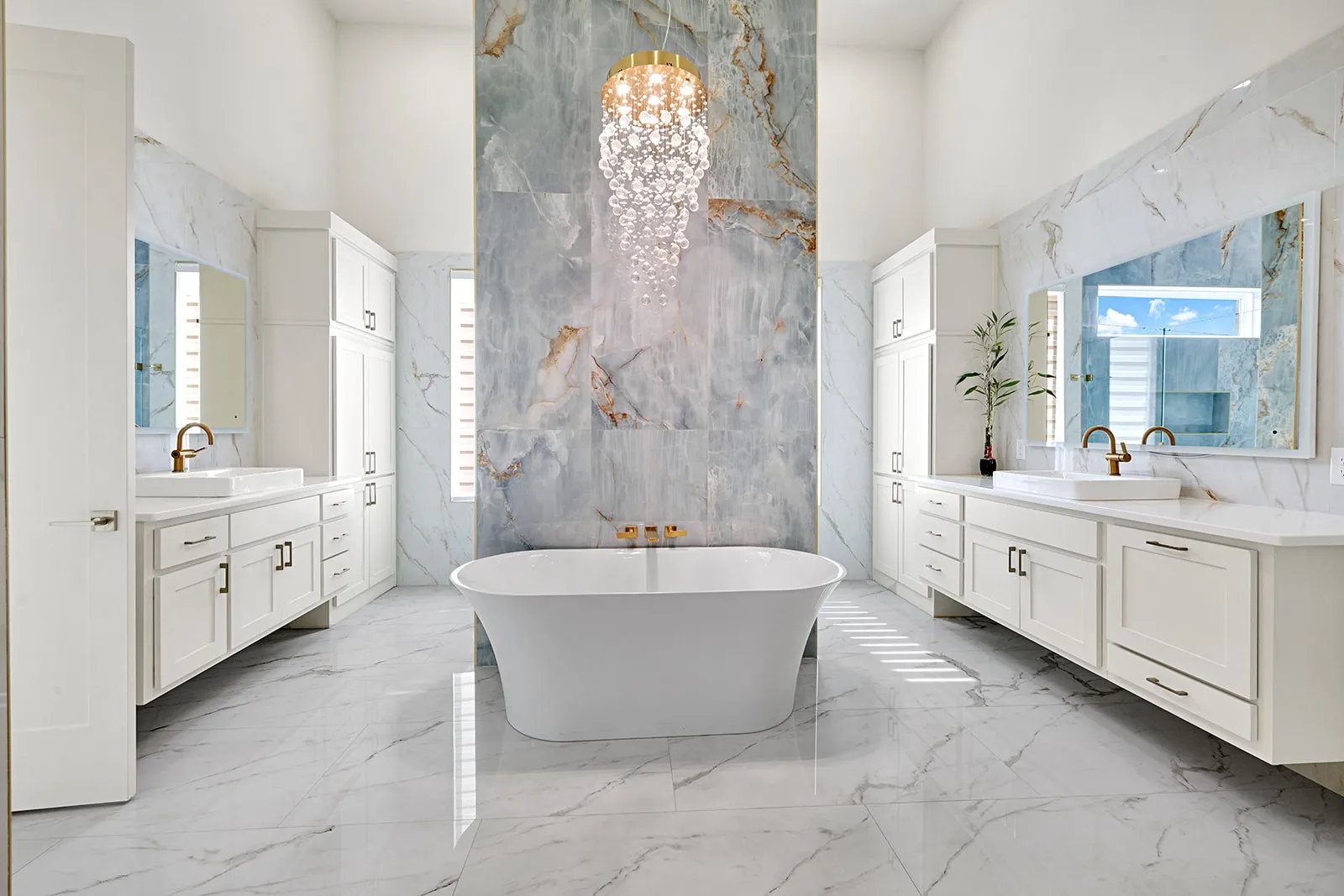 Spacious bathroom featuring a freestanding soaking tub, dual vanity areas with white cabinetry and vessel sinks, a vaulted ceiling, and floor-to-ceiling marbled accent wall