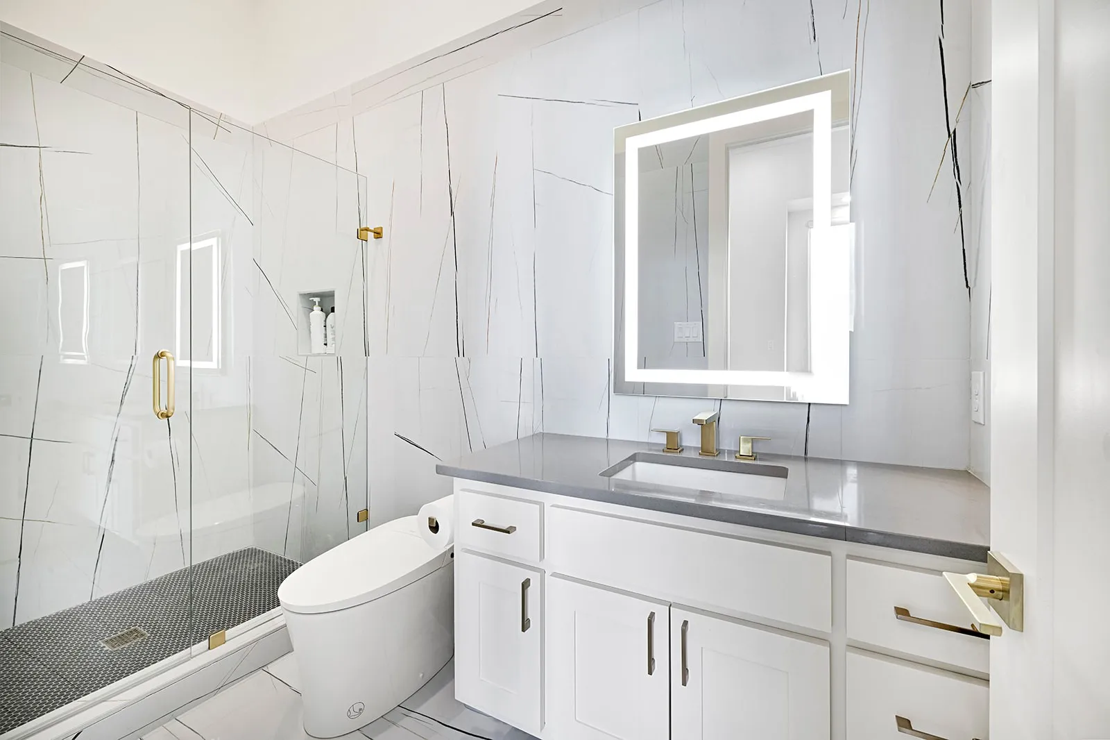 Modern bathroom featuring a frameless glass shower enclosure with gold-tone hardware, a wall-mounted vanity with a light-colored countertop, and an illuminated mirror