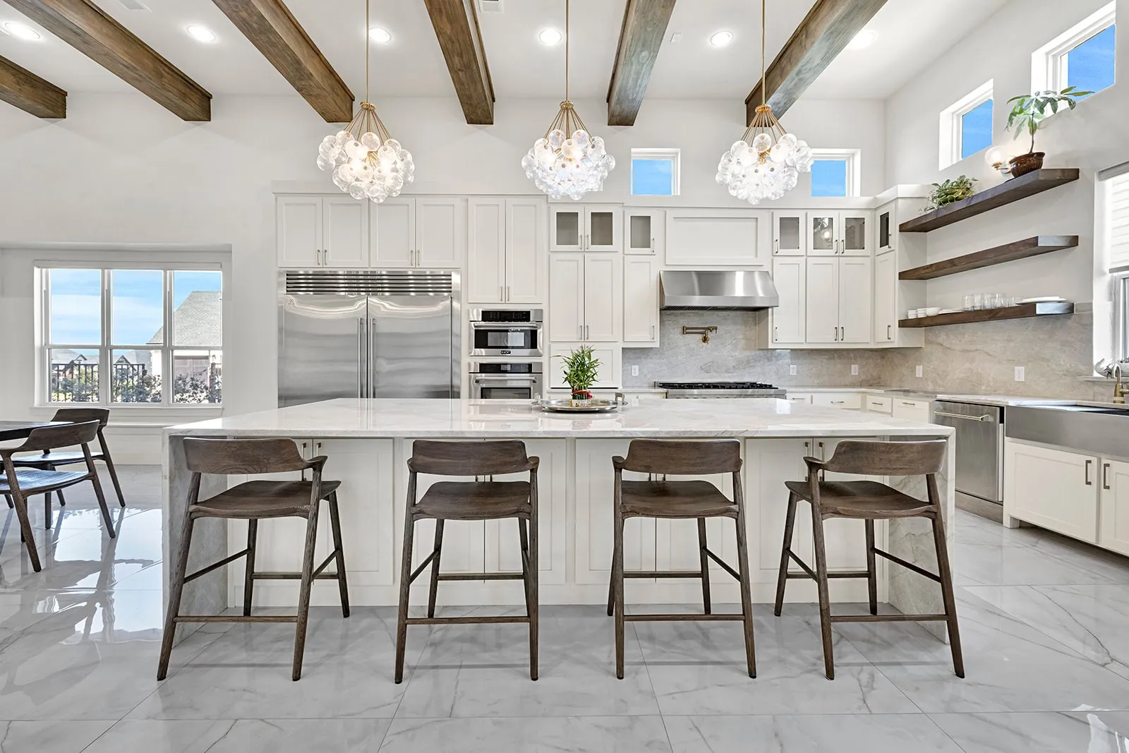 Spacious kitchen featuring high ceilings with exposed wood beams, a large central island with seating, stainless steel appliances, white cabinetry, and light-toned stone countertops