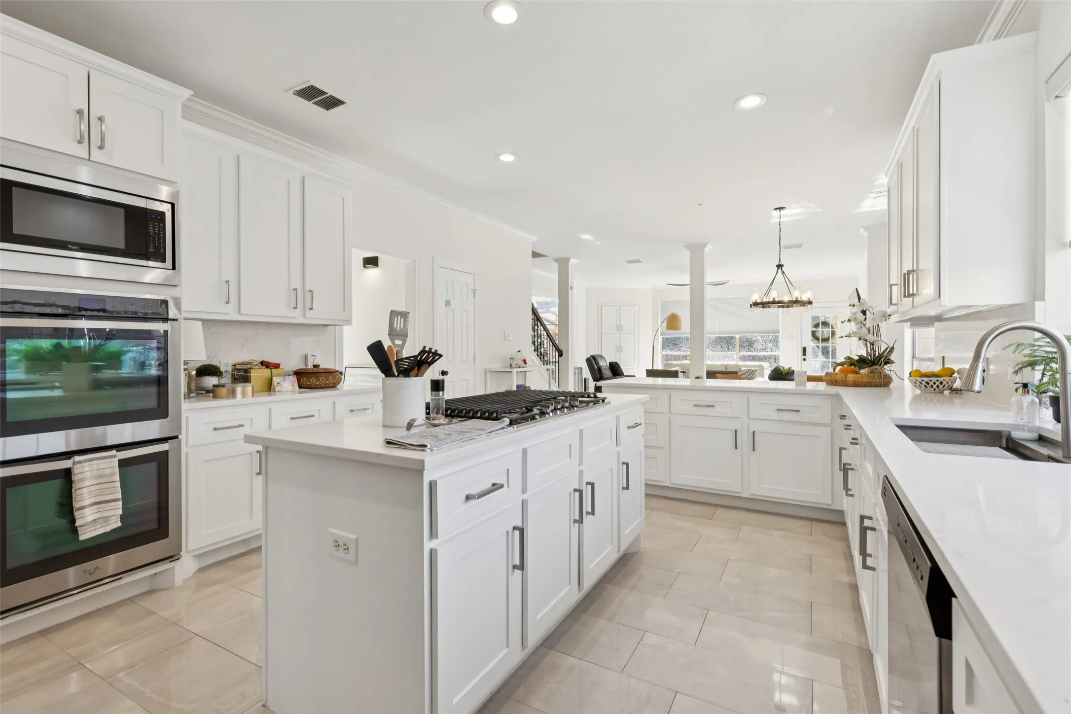 Bright kitchen featuring white cabinetry, light-toned countertops, stainless steel appliances including a built-in microwave and double oven, a central island with an integrated cooktop, and large format tile flooring