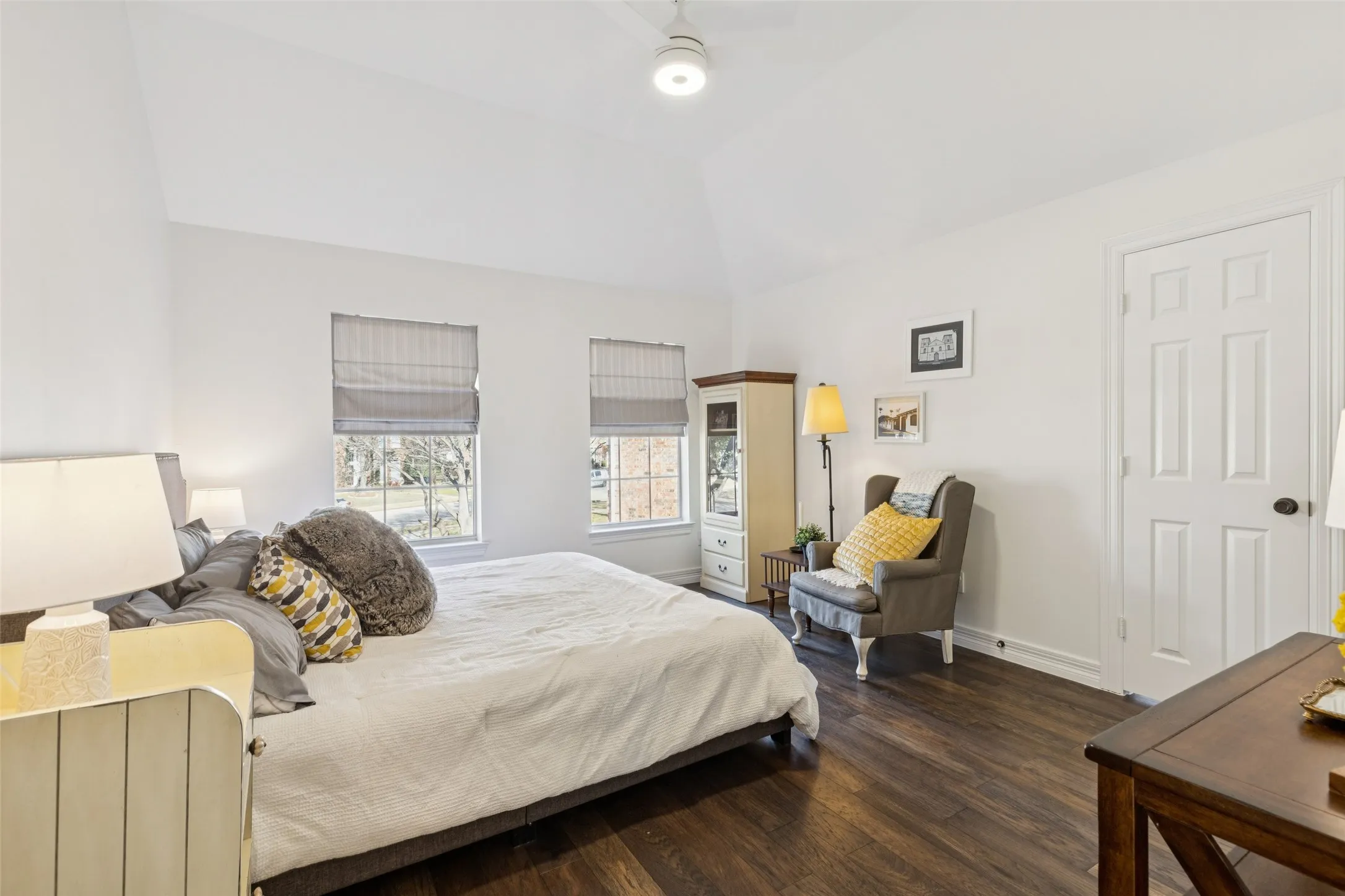 Vaulted ceiling room featuring dark wood-finish flooring, two windows with Roman shades, a ceiling fan, and a paneled interior door