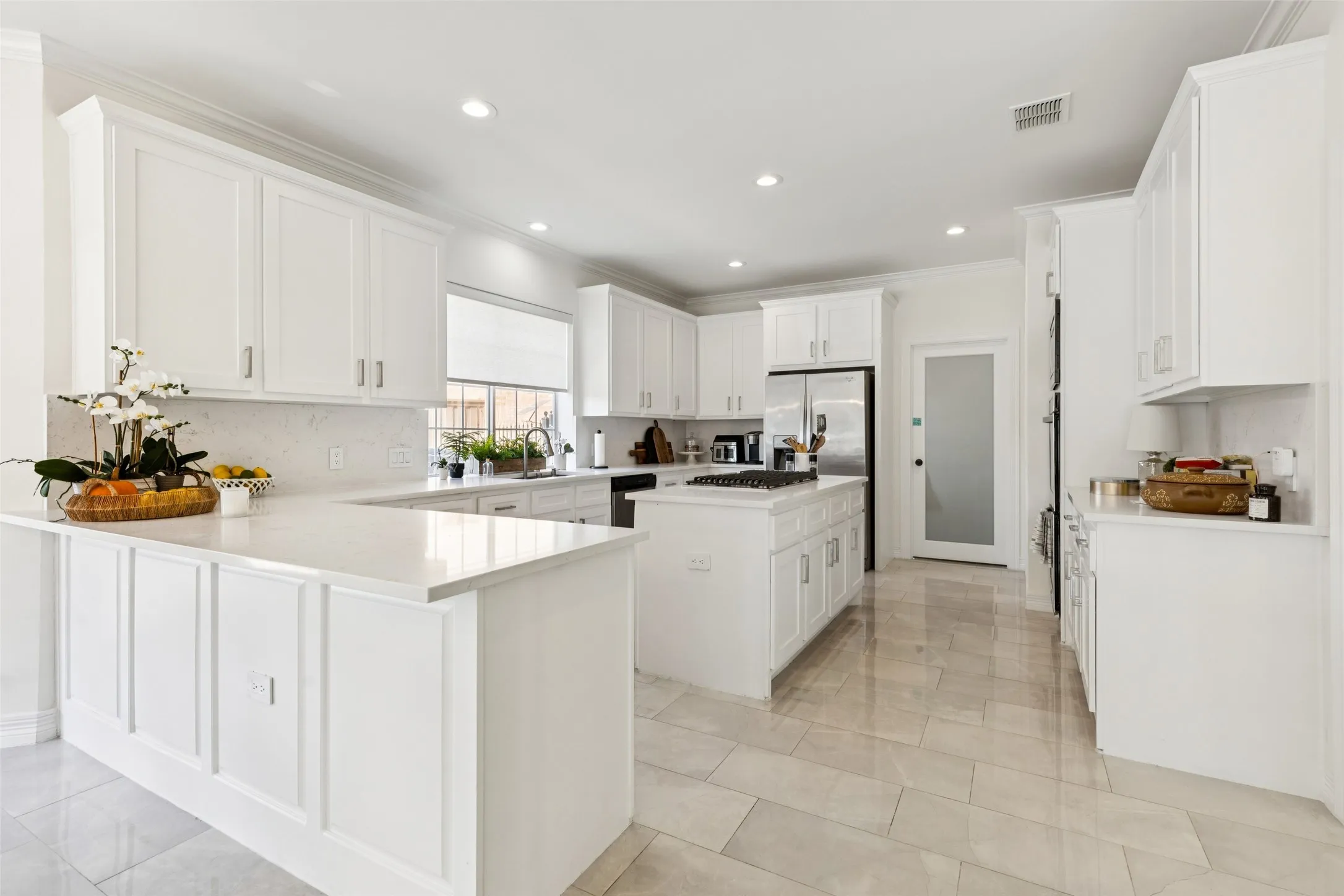 Spacious kitchen featuring extensive white cabinetry, light-toned tiled flooring, white countertops, a central island with a built-in cooktop, and stainless steel appliances