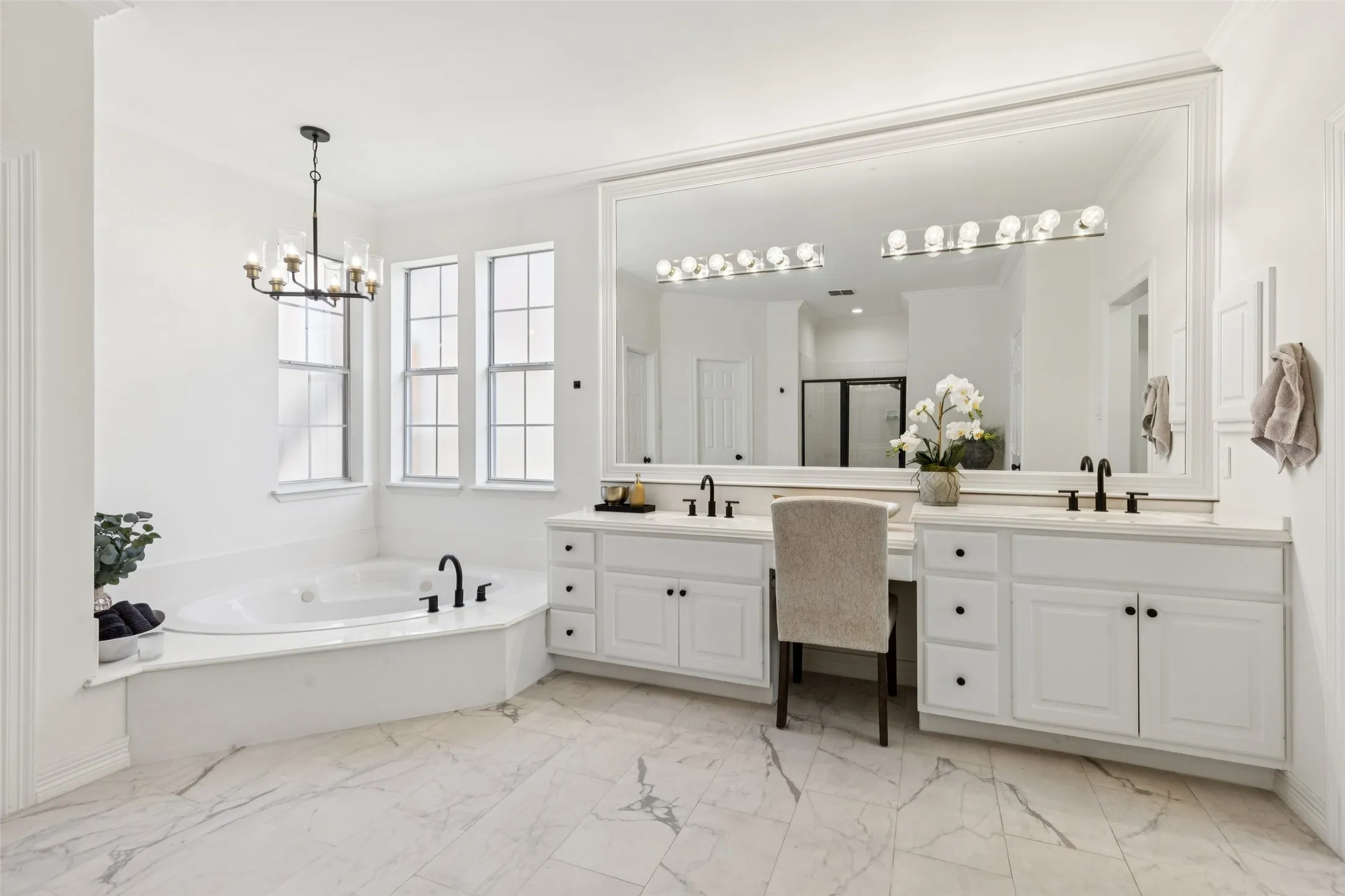 Spacious bathroom featuring a built-in corner soaking tub, dual vanities with white cabinetry, a large vanity mirror, and marble-look tile flooring