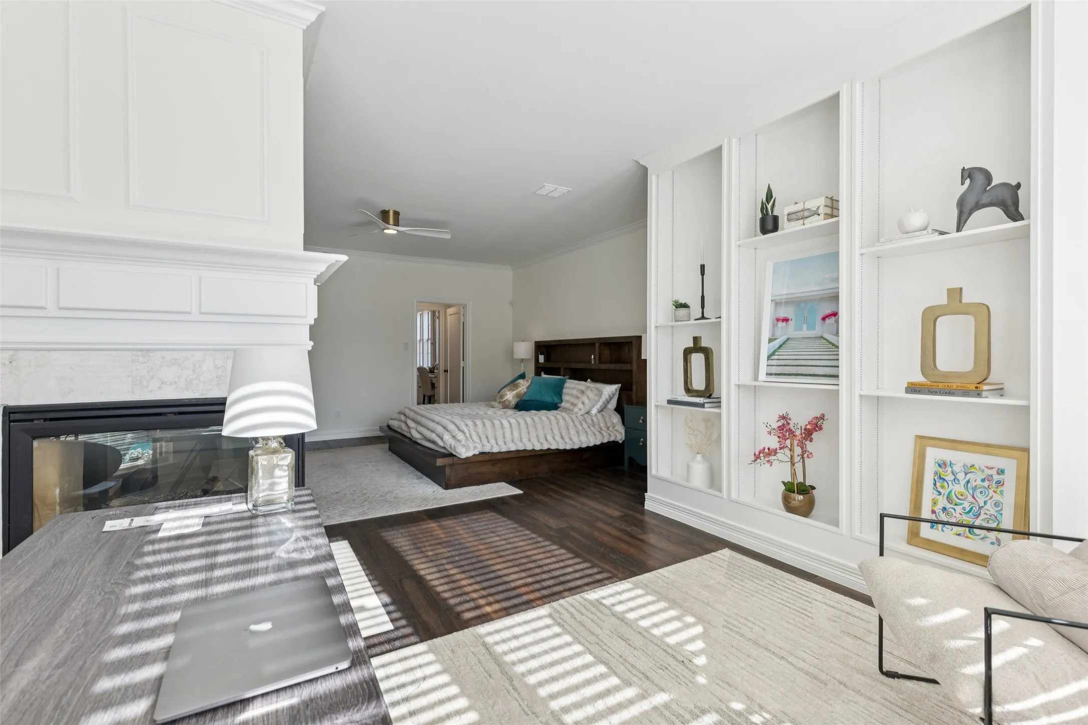 Spacious room featuring a white paneled fireplace with a dark-faced insert, built-in shelving, dark wood-finish flooring, light-toned area rug, and a ceiling fan with gold accents