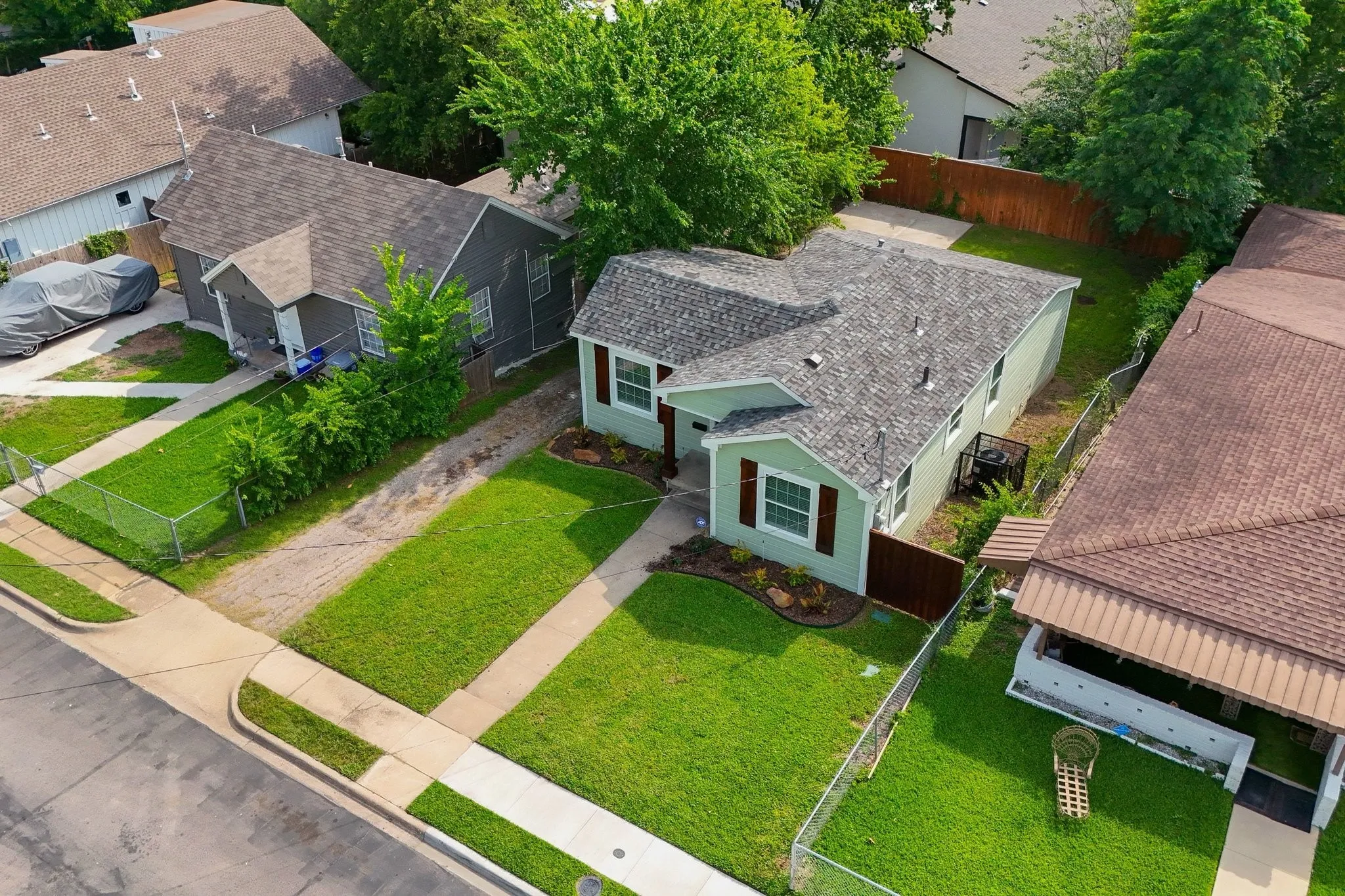 Light green siding home featuring a dark shingle roof, brown window shutters, and a well-maintained front lawn with established landscaping