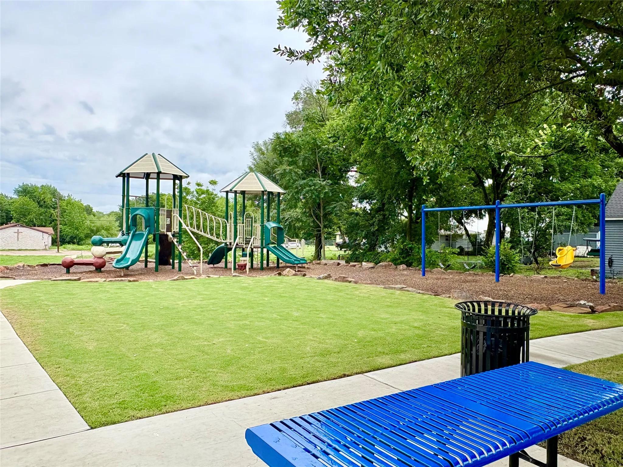 Recreational area featuring a multi-level play structure with slides and climbing elements, a swing set, a manicured lawn, mature trees, and a concrete pathway