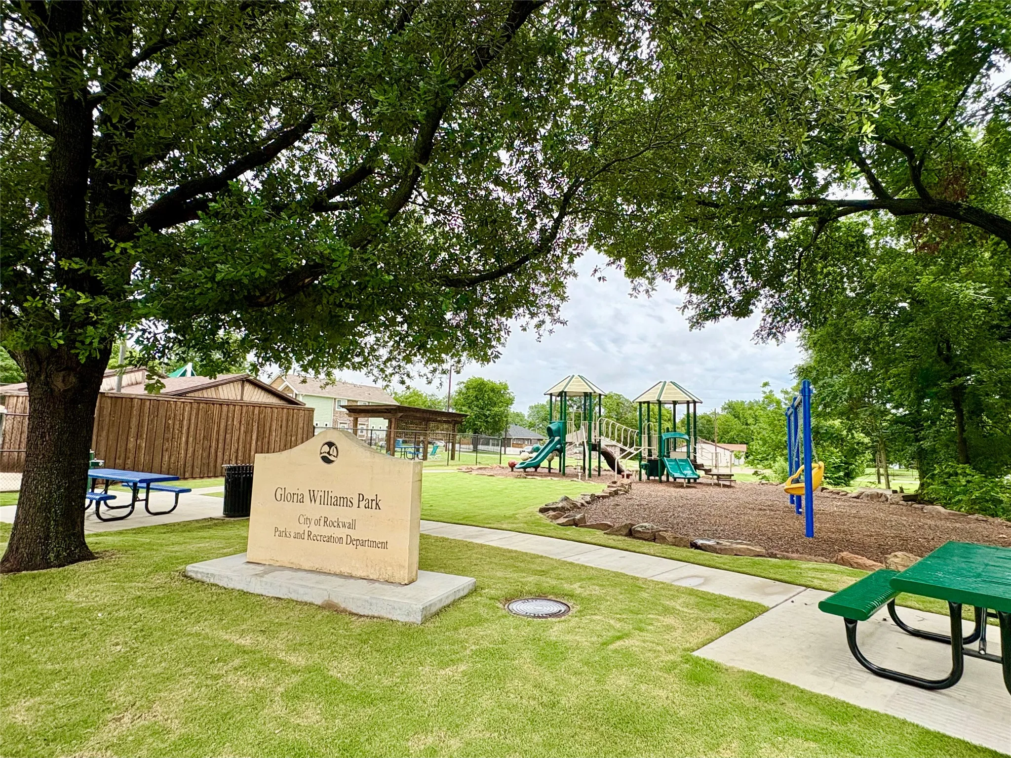 Expansive green space featuring a large shade tree, a concrete park sign, a paved pathway, and a playground with slides and swings