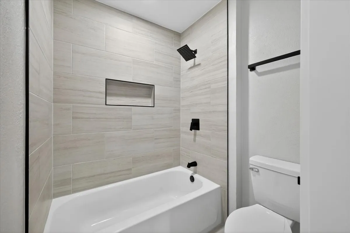 Bathtub and shower combination featuring light-toned tile surround, matte black shower fixtures, a recessed shower niche, and a white porcelain toilet