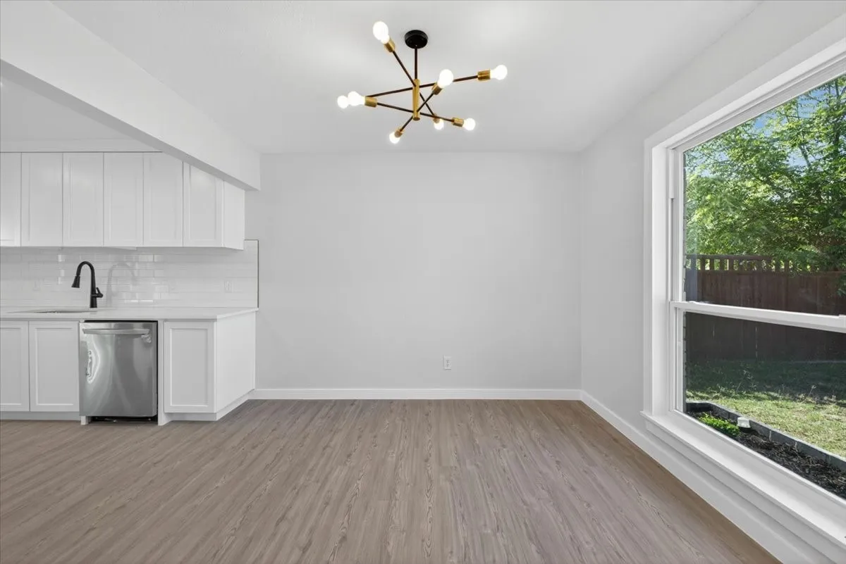 Open-concept living space featuring wood-finish flooring, white cabinetry, a stainless steel dishwasher, a modern sputnik chandelier, and a large window overlooking the backyard